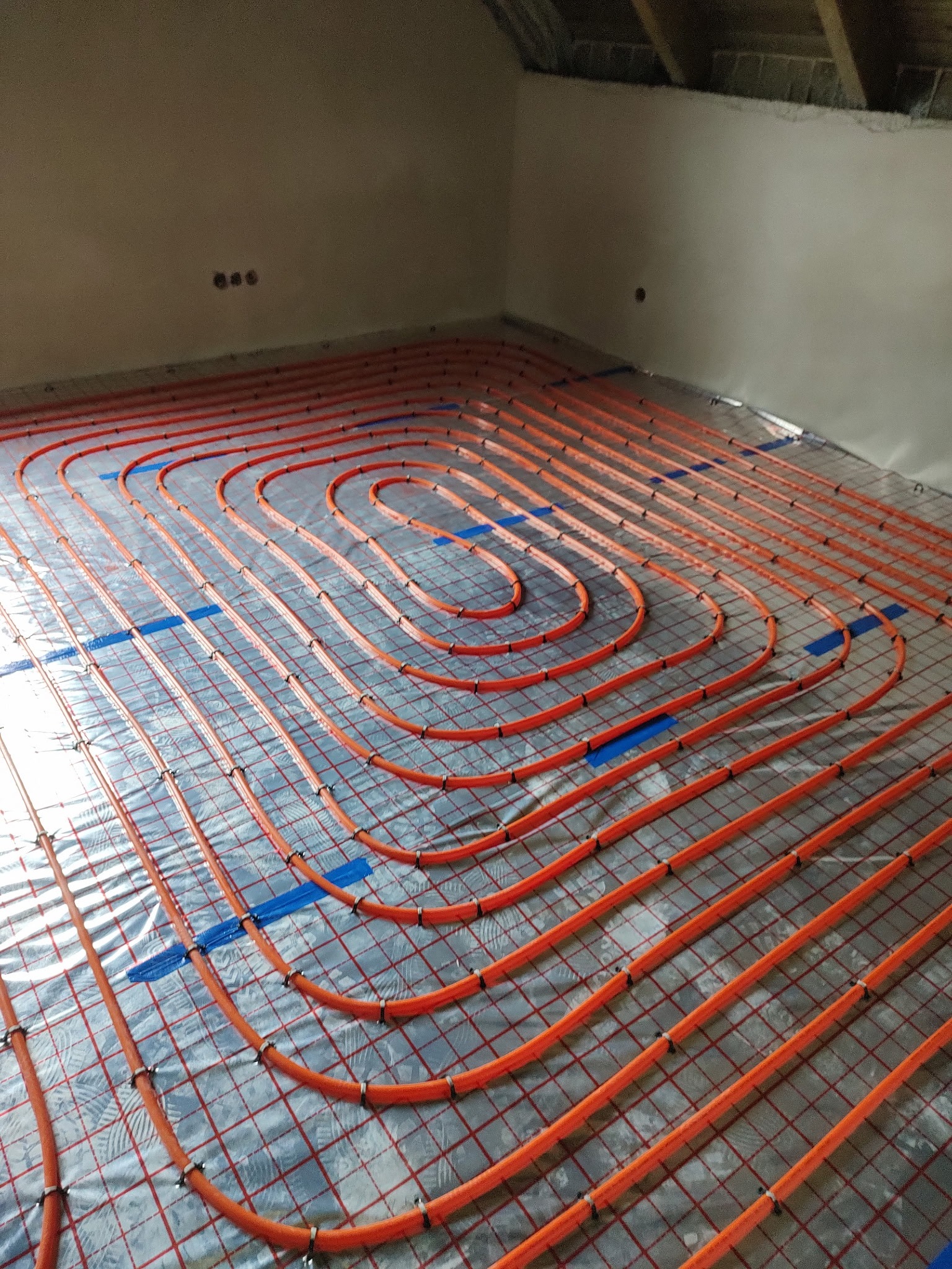 Pomarańczowe rury ogrzewania podłogowego ułożone na srebrnej folii z czerwoną siatką, przymocowane niebieską taśmą, widok z góry na surową ścianę i poddasze.