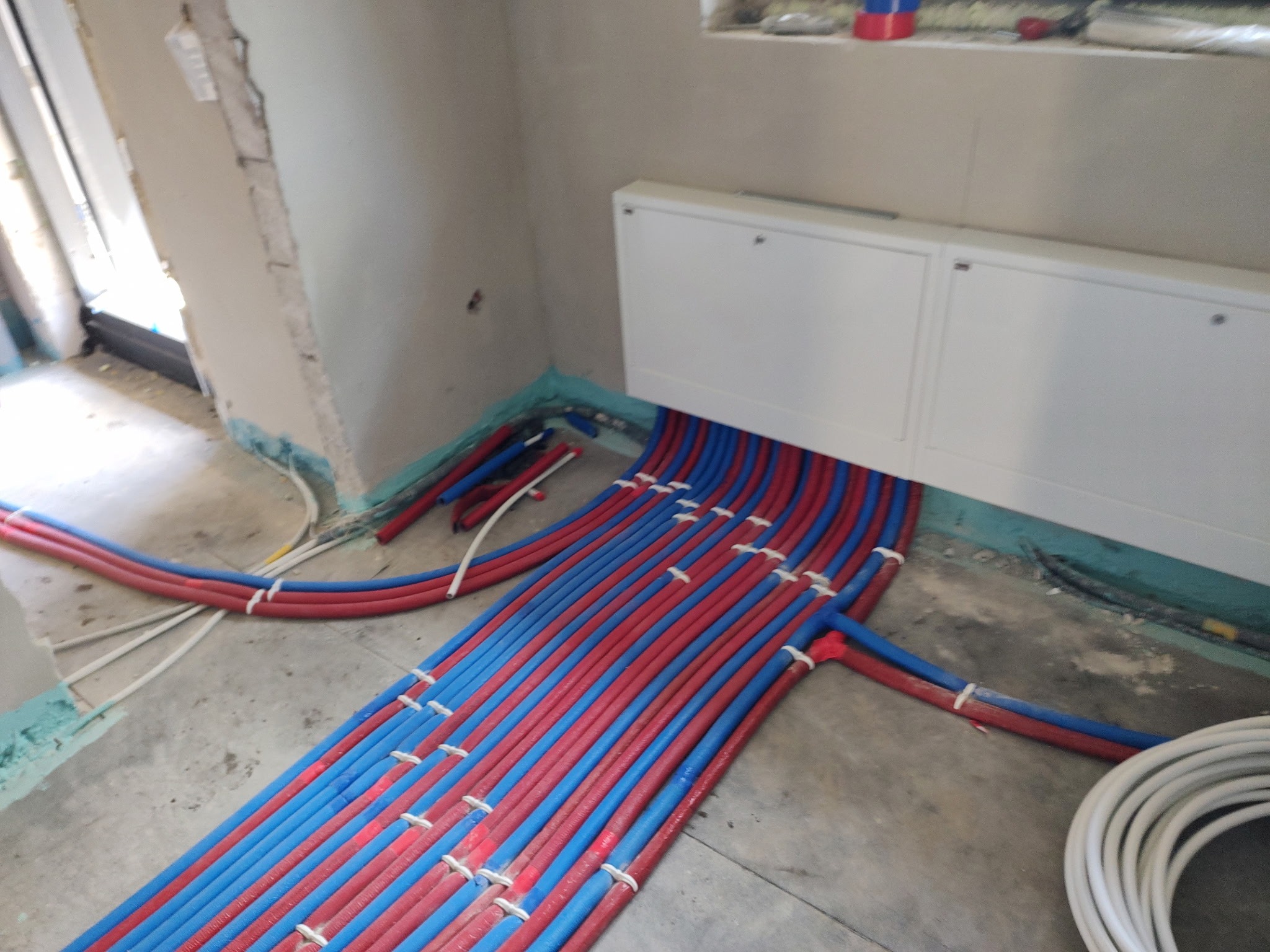 Instalacja ogrzewania podłogowego z rurami w kolorze czerwonym i niebieskim, ułożonymi równolegle na szarym betonie, przed wylaniem posadzki. Widoczne białe szafki.