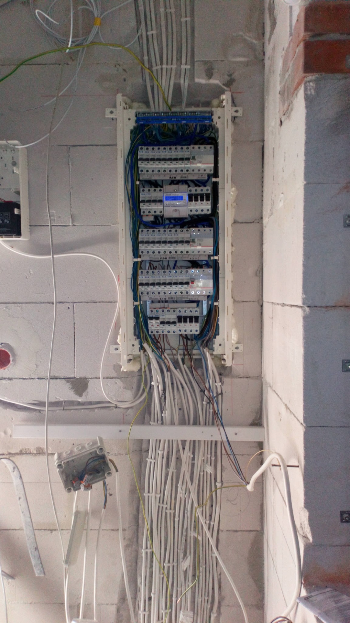 Rozdzielnica elektryczna z licznymi przewodami w surowym wnętrzu. Widoczne zabezpieczenia i podłączenia, instalacja w trakcie montażu na ścianie z betonu komórkowego.