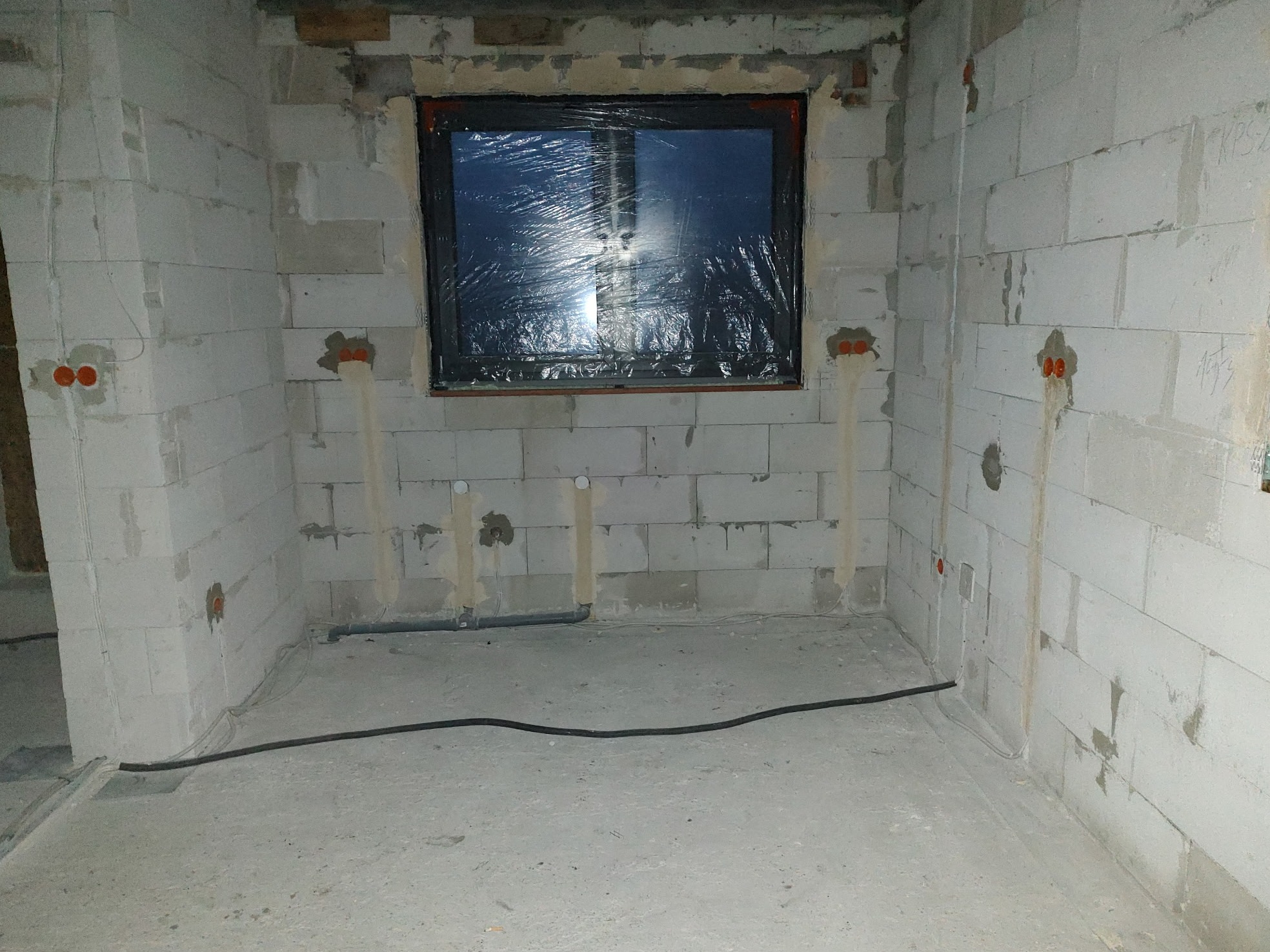 Surowy stan: ściany z betonu komórkowego z instalacją elektryczną i rurami, okno zabezpieczone folią. W tle widoczne przewody na podłodze.