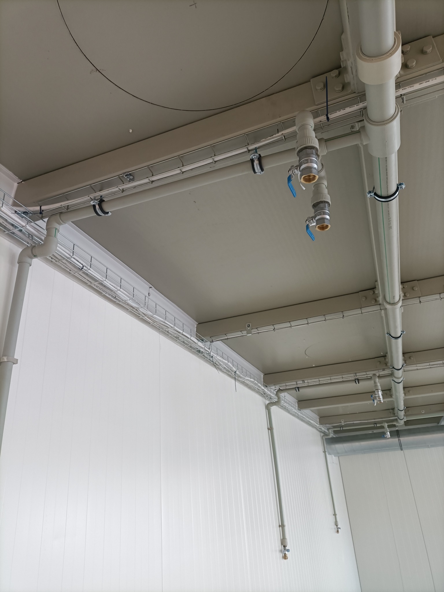 Szara instalacja rurowa z zaworami, zamocowana pod sufitem w pomieszczeniu o białych ścianach. Widoczne kable w metalowych korytkach.