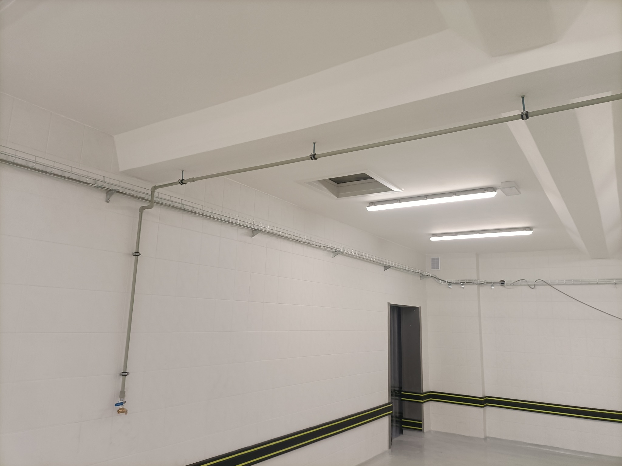 Białe pomieszczenie z instalacją rur pod sufitem i przy ścianie, zabezpieczenia na dole ścian, oświetlenie sufitowe i kratka wentylacyjna.