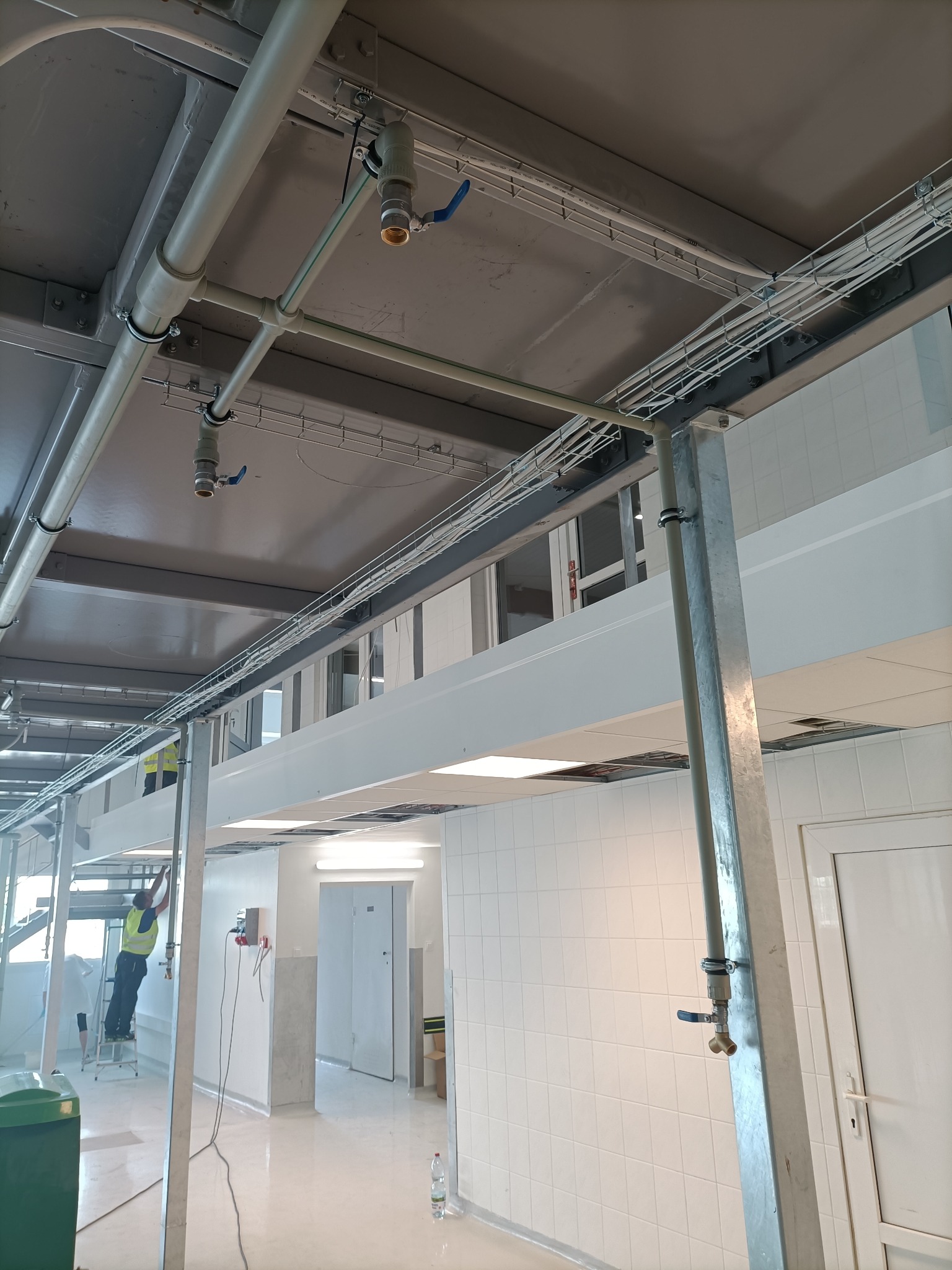 Instalacja rur i kabli pod sufitem w pomieszczeniu z białymi płytkami. Widoczny pracownik na drabinie i elementy instalacji.