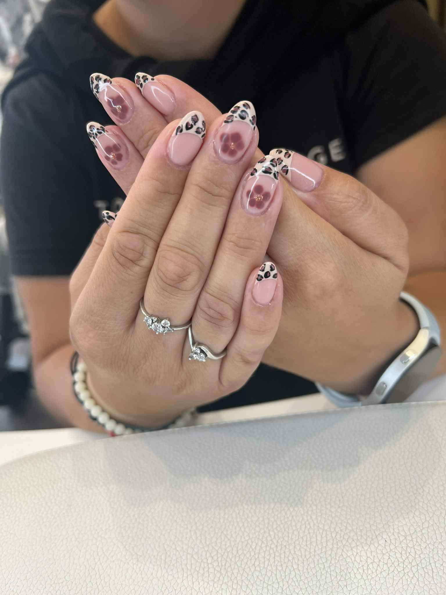 Dłonie kobiety z manicure hybrydowym: beżowe paznokcie z panterkowym wzorem na końcach i kwiatowym akcentem na dwóch paznokciach. Widoczny pierścionek i bransoletka.