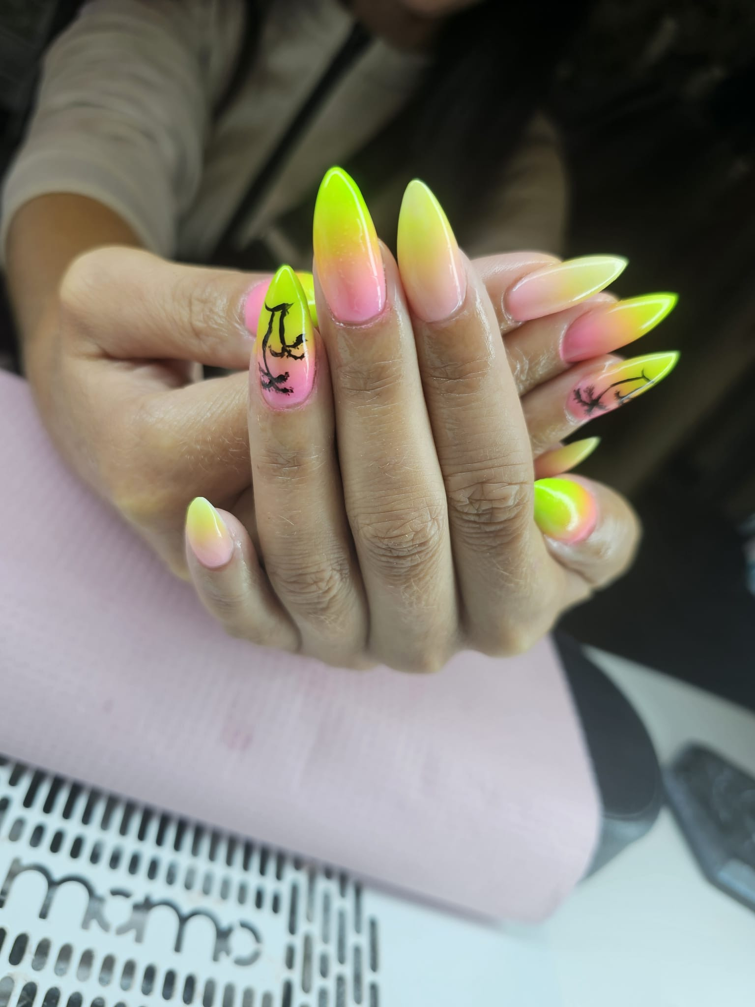 Dłonie z neonowymi, ombre paznokciami w odcieniach różu i żółci, zdobione motywem palmy. Ostry kształt paznokci. Manicure wykonany na stole z lampą UV.