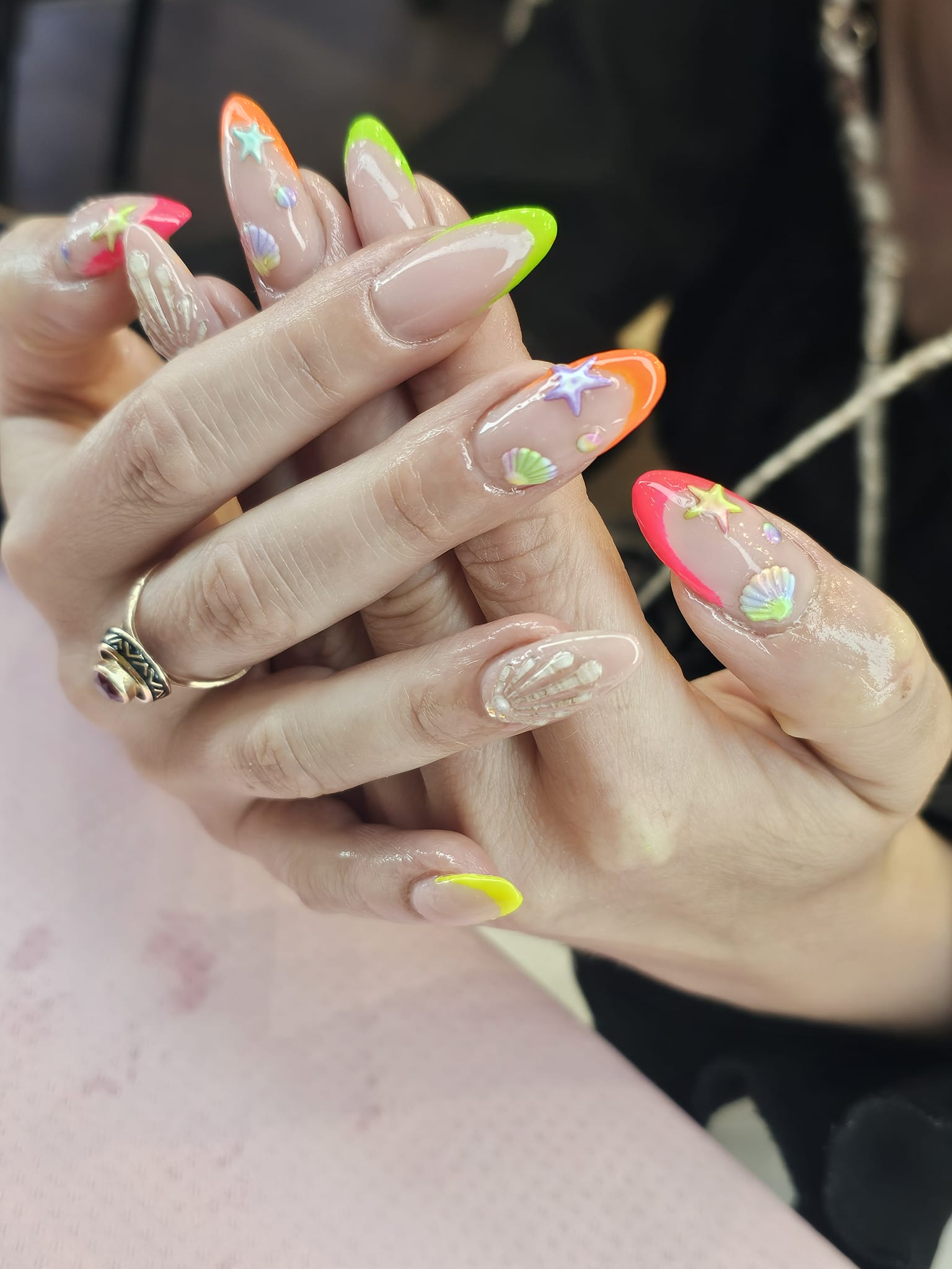 Dłonie z długimi paznokciami, zdobionymi neonowymi końcówkami i motywami morskimi (muszelki, gwiazdki). Pierścionek na palcu. Jasne tło.