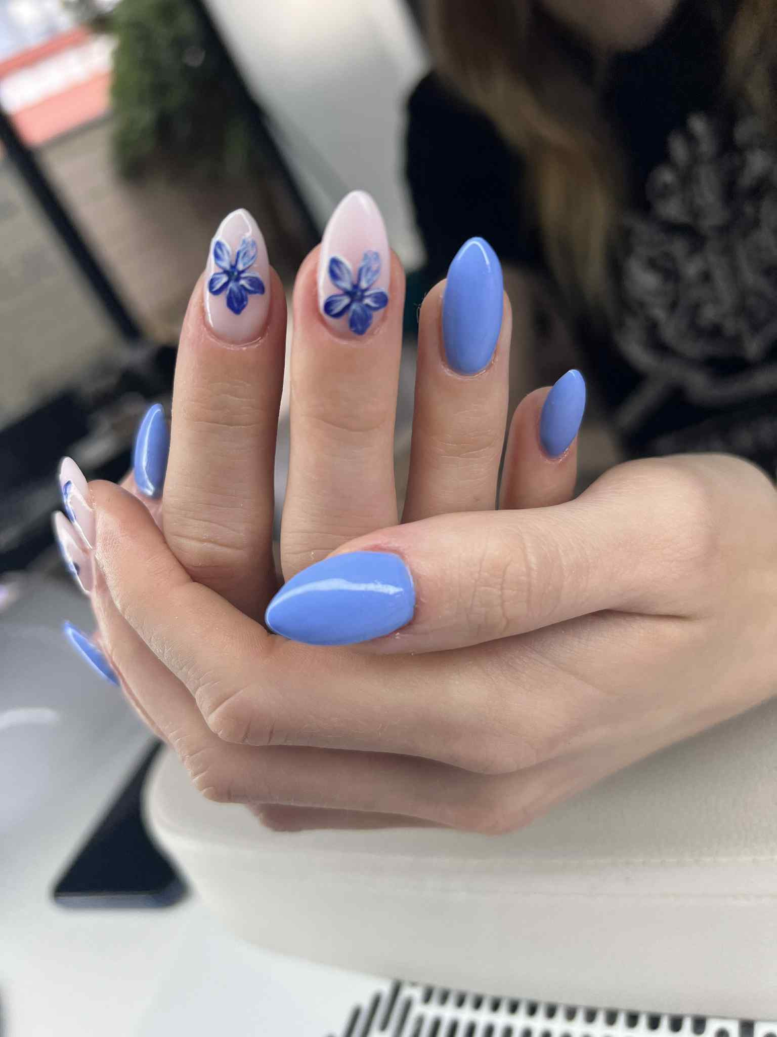 Dłonie z niebieskim manicure hybrydowym, dwa paznokcie z kwiatowym wzorem. Migdałowy kształt paznokci, jasna karnacja dłoni, delikatne światło.