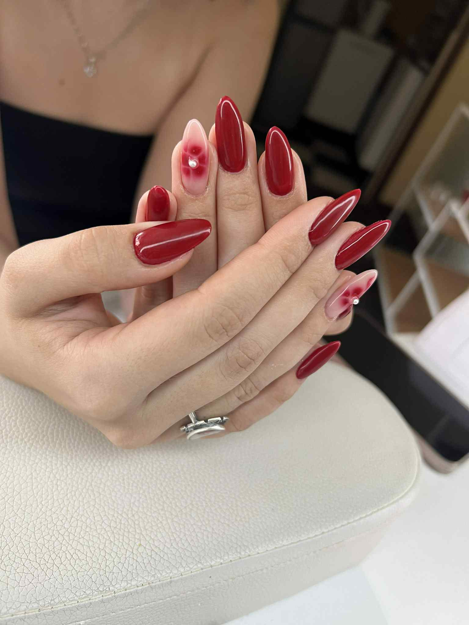 Dłonie z czerwonym manicure hybrydowym, kształt szpiczasty. Jeden paznokieć z efektem ombre i kwiatowym zdobieniem 3D. Pierścionek na palcu.