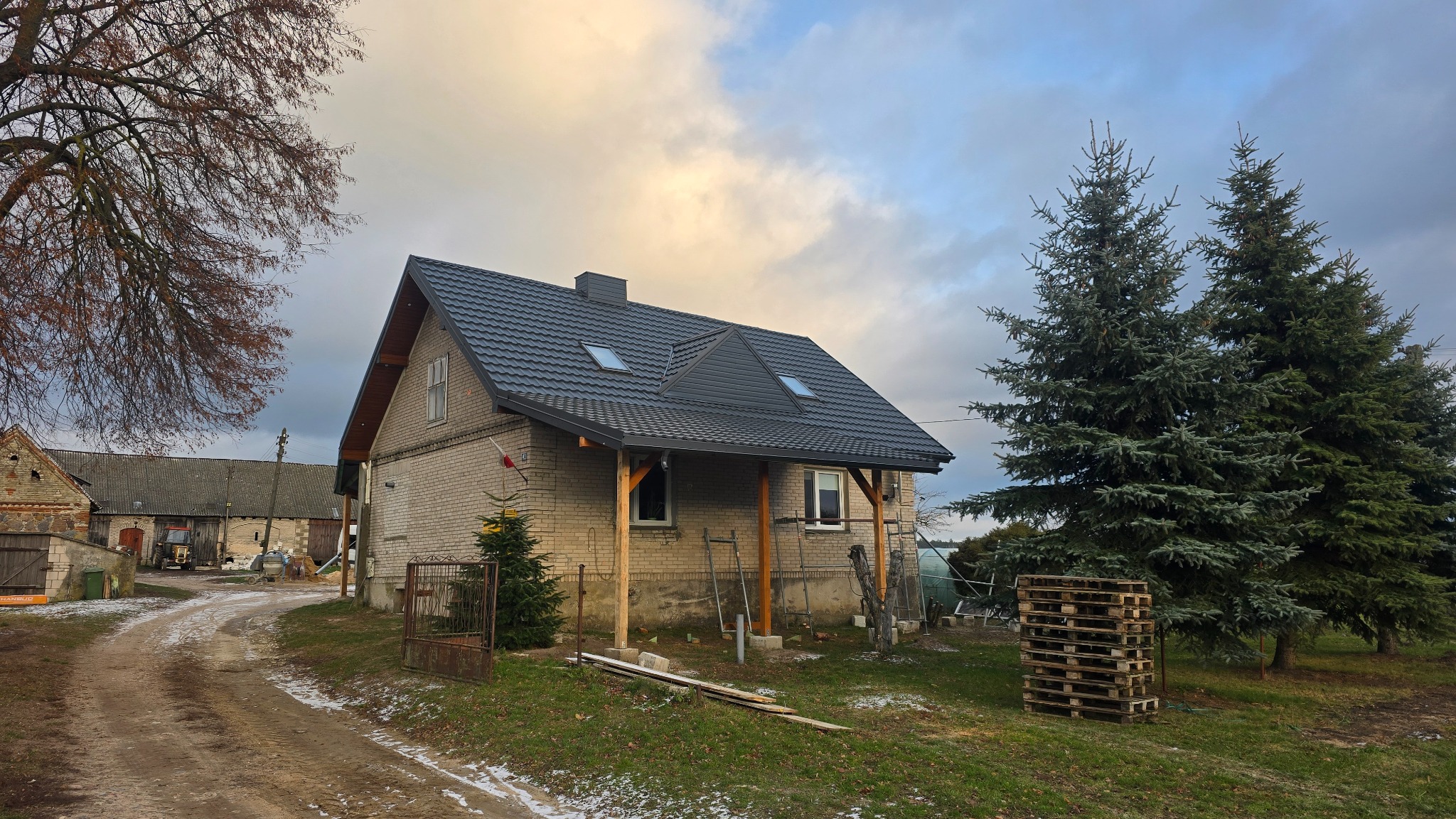 Dom z nowym, grafitowym dachem z blachodachówki i oknami dachowymi. Widoczne rusztowania i palety, sugerujące prace wykończeniowe. Pochmurne niebo w tle.