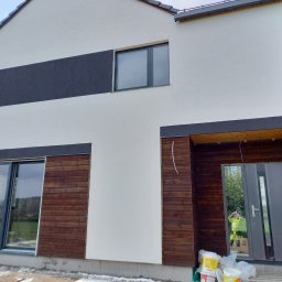 Jerzy Zbigniew Bodak - Nowoczesny dom z białą elewacją i drewnianymi akcentami. Widoczne okna, drzwi wejściowe i fragment osoby w odbiciu. Częściowo wykończona elewacja.