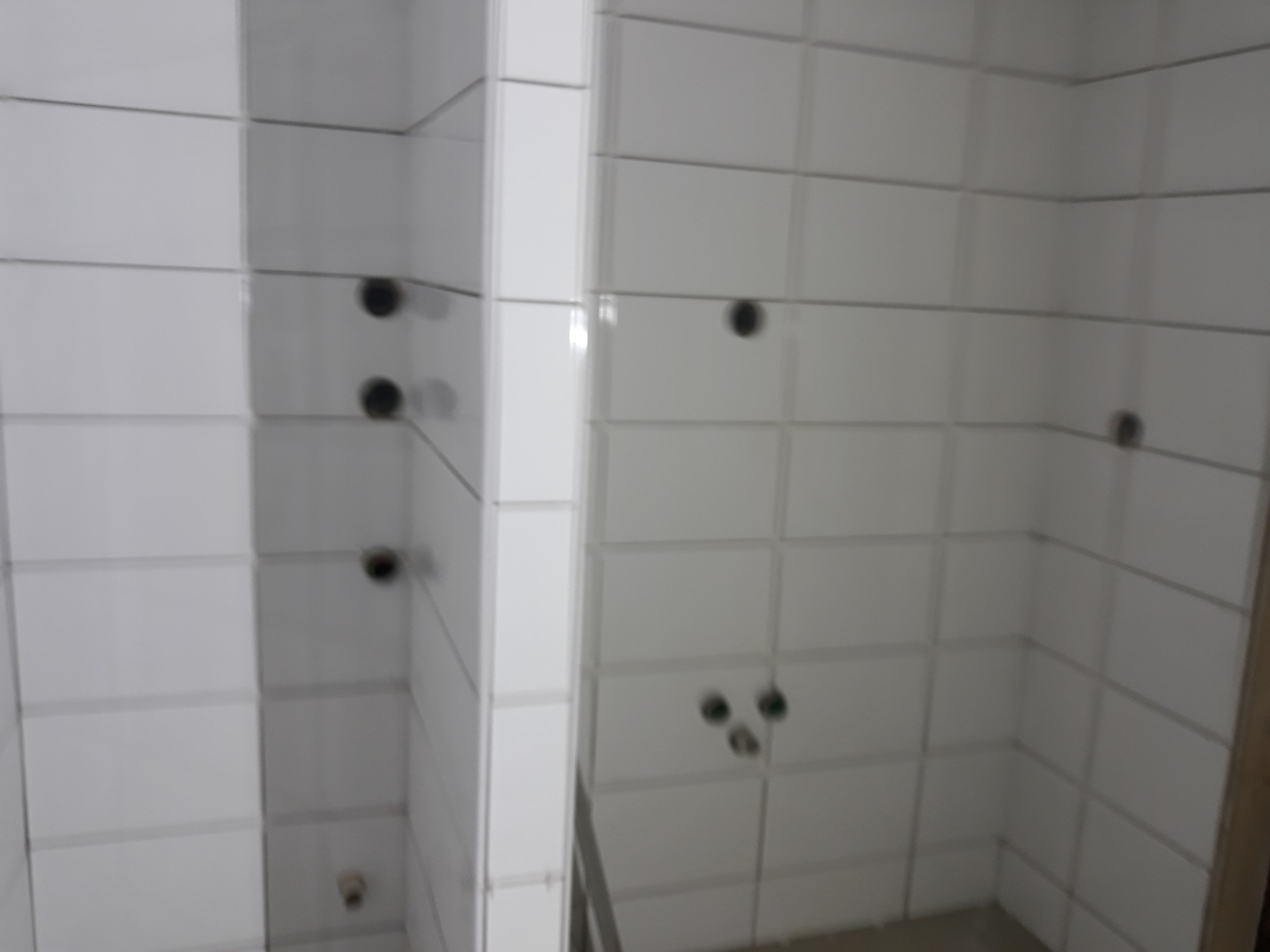 Wykończona ściana łazienki z białymi, prostokątnymi płytkami i otworami na armaturę. Widok na narożnik z widocznymi fugami i punktami instalacji.