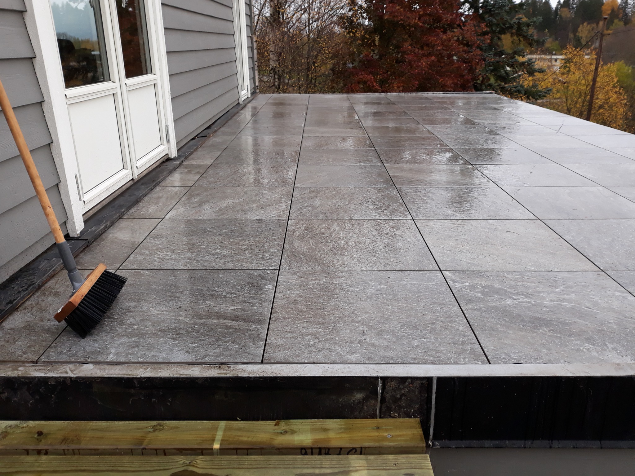 Ułożona, szara, kwadratowa terakota na tarasie przylegającym do domu z szarą elewacją. Widoczna szczotka do zamiatania. Tło stanowią drzewa z jesiennymi liśćmi.