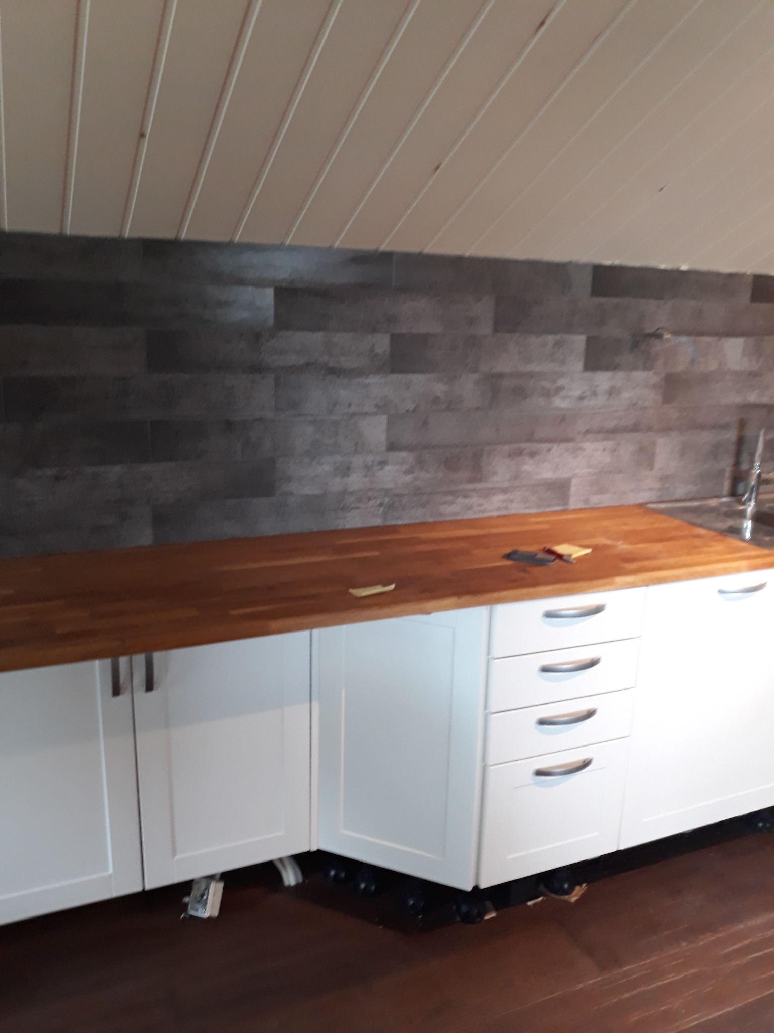 Nowoczesna zabudowa kuchenna z białymi szafkami i drewnianym blatem, uzupełniona o szarą, ceglaną ścianę. Widoczny fragment poddasza z drewnianą boazerią.