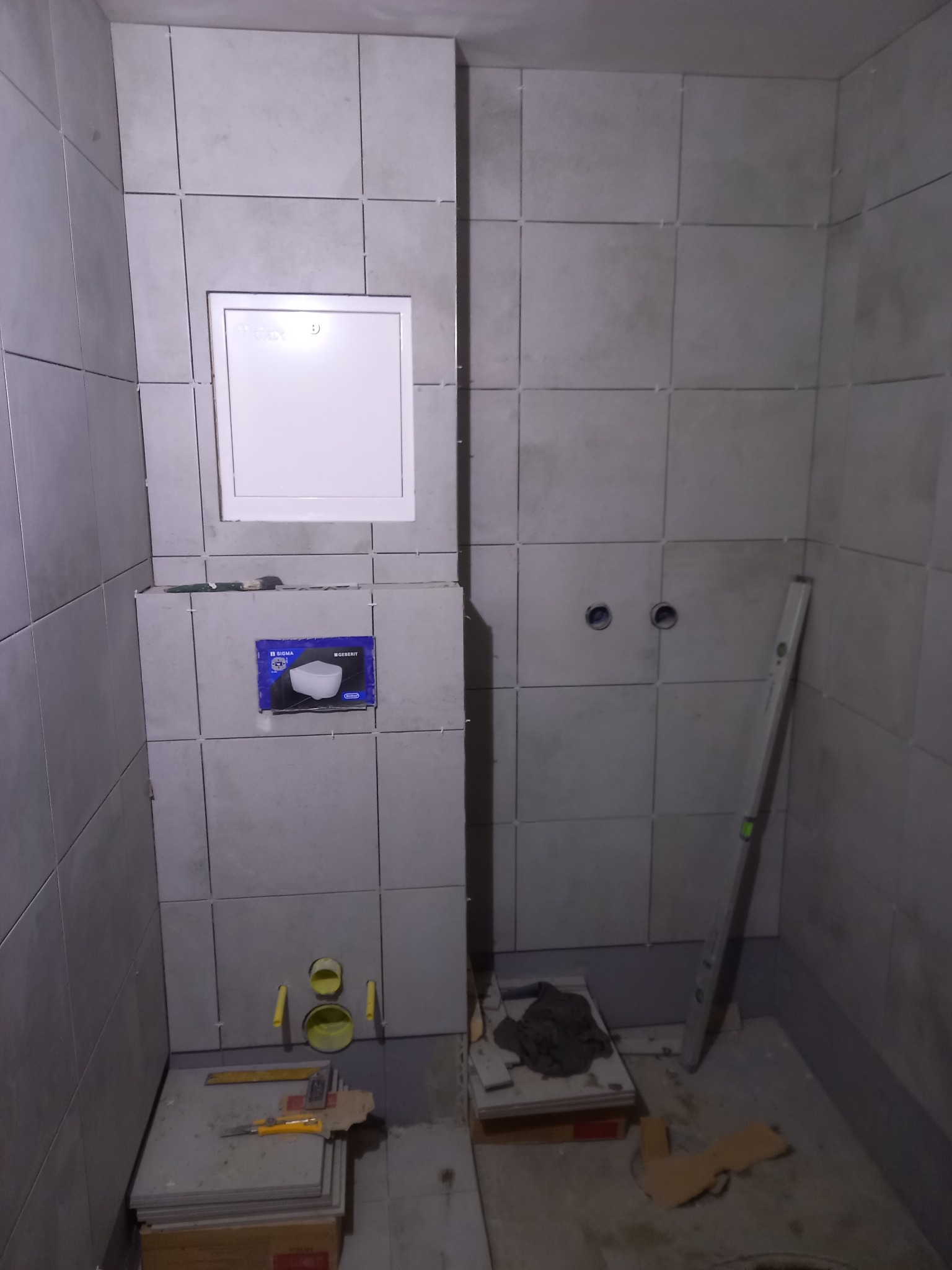 Ściana łazienki w trakcie układania płytek ceramicznych w odcieniach szarości. Widoczny otwór rewizyjny, poziomica i narzędzia glazurnicze. Etap prac wykończeniowych.