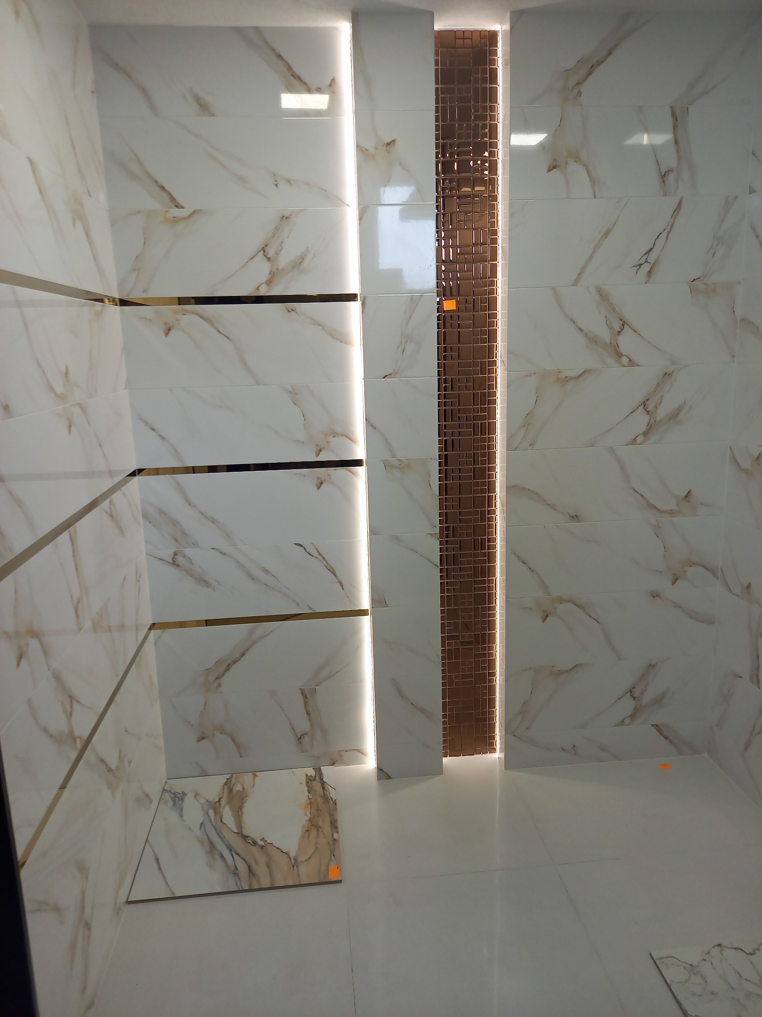 Wykończona łazienka z białymi płytkami imitującymi marmur, złotymi akcentami i dekoracyjną mozaiką w pionowym pasie z podświetleniem LED. Nowoczesny design.