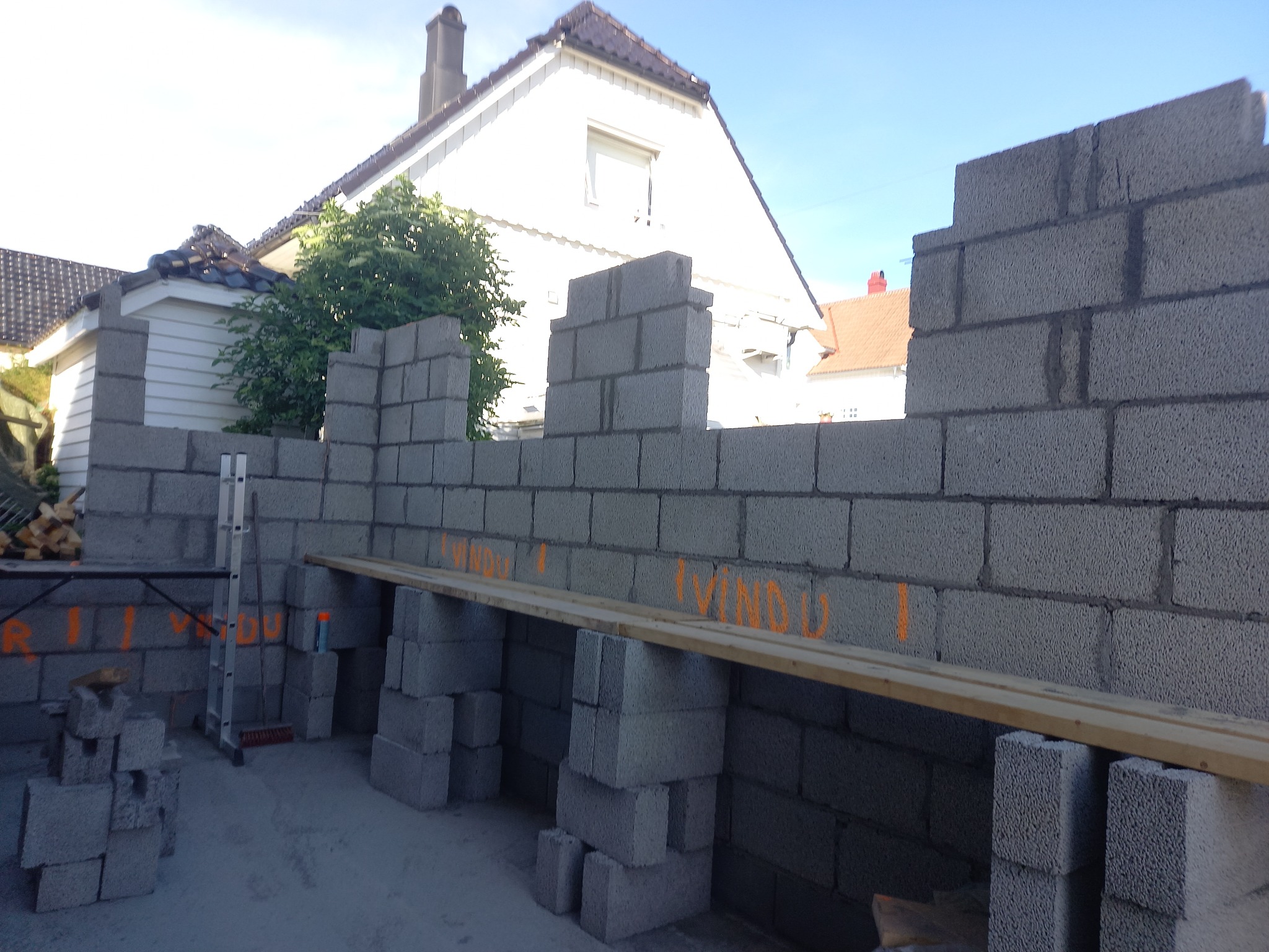 Wznoszenie ściany z szarego bloczka betonowego na budowie domu jednorodzinnego. Widoczne rusztowanie i zapas bloczków. Na ścianie pomarańczowe oznaczenia.