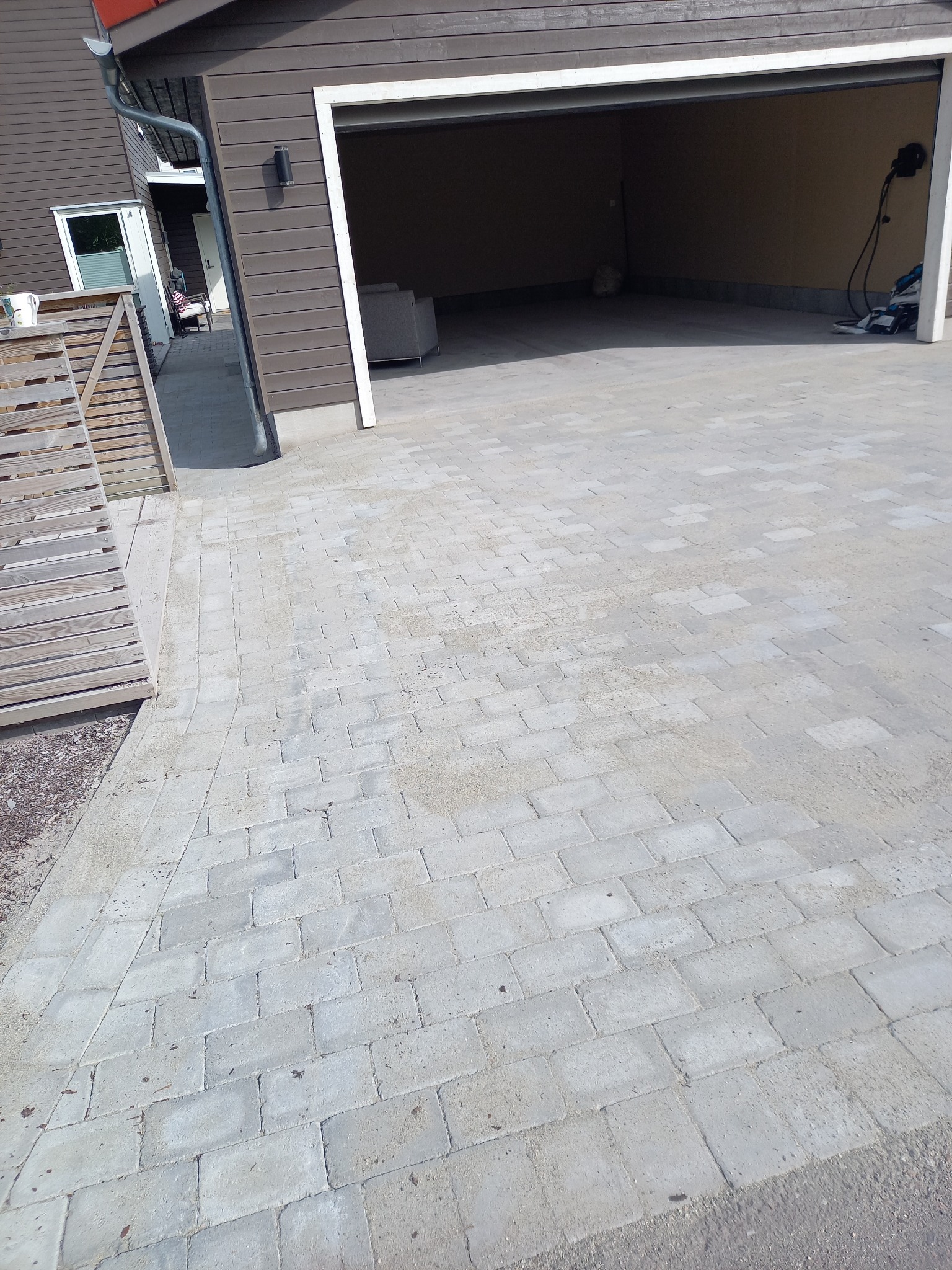 Ułożona szara kostka brukowa przed garażem z brązową elewacją. Widoczny fragment drewnianego płotu i drzwi wejściowe do domu. Jasne, naturalne światło.