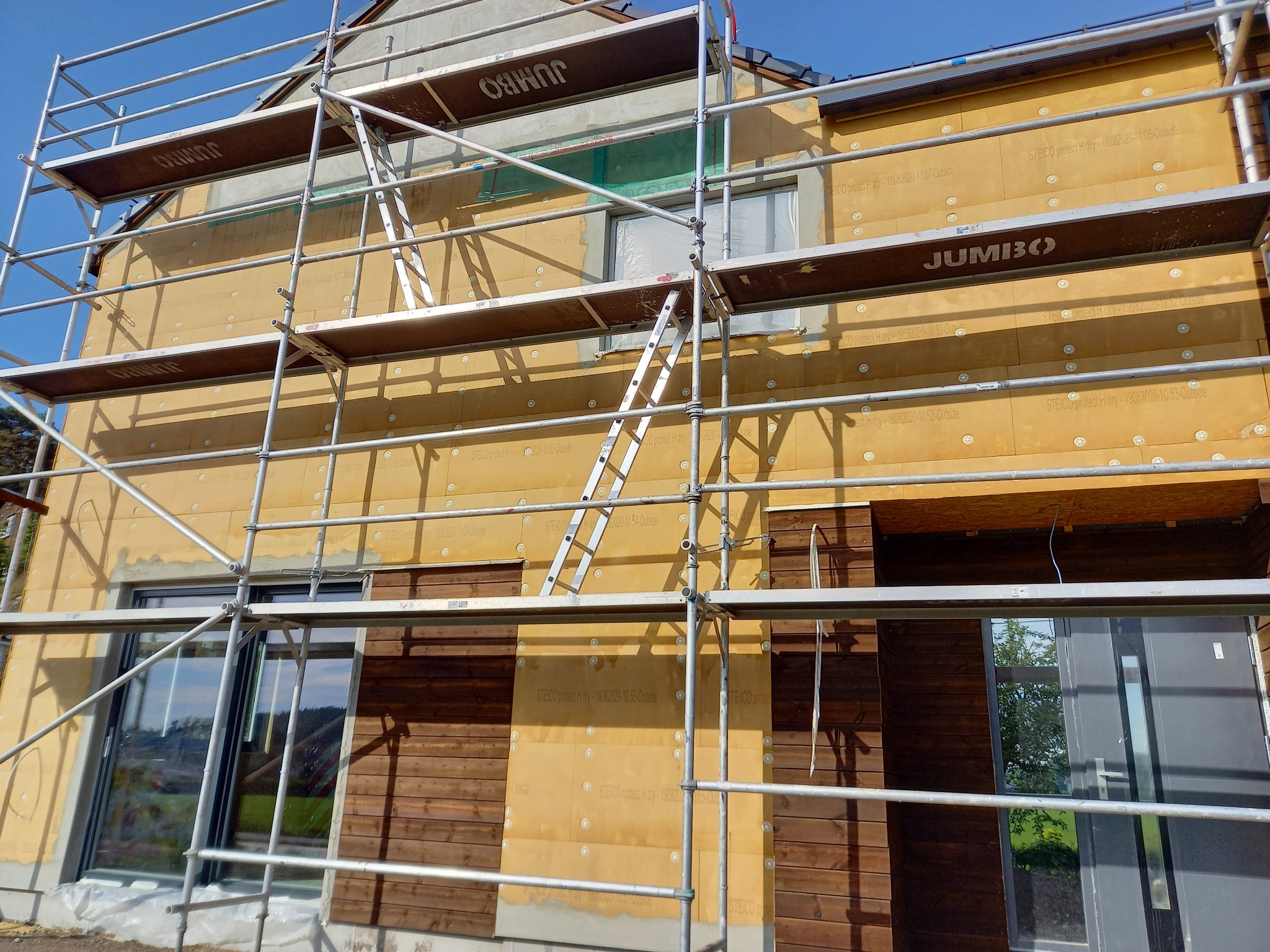 Ocieplanie elewacji budynku z użyciem rusztowania. Widoczne płyty izolacyjne STEICO protect H dry. Część elewacji wykończona drewnem. Słoneczny dzień.