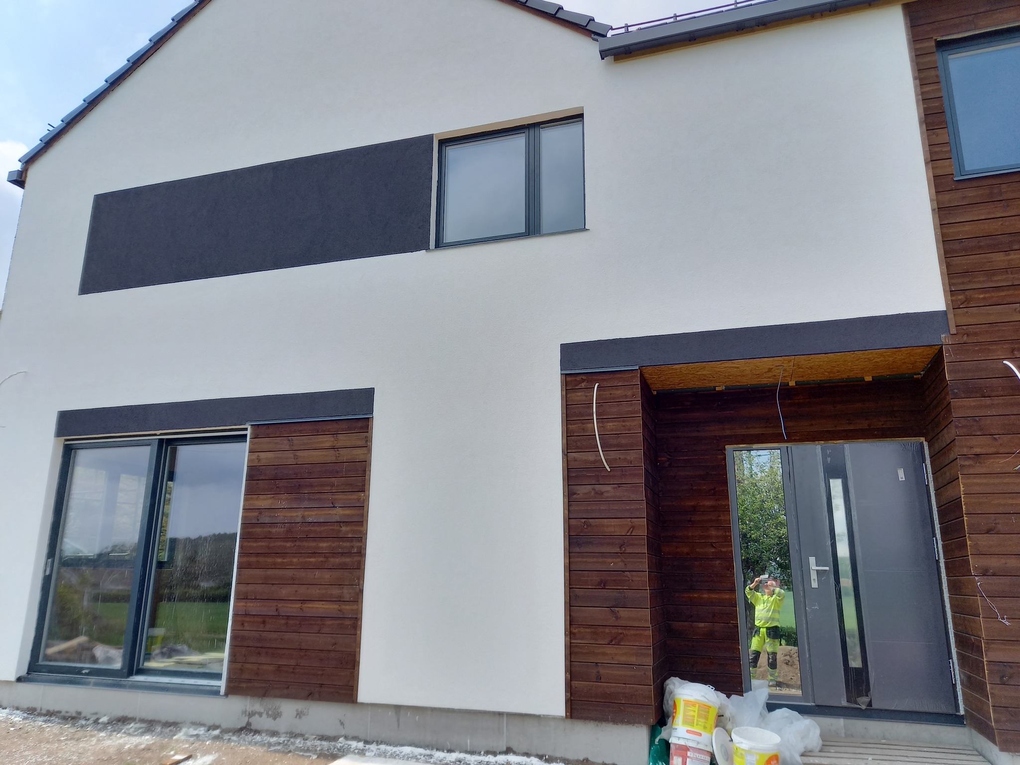 Nowoczesny dom z białą elewacją i drewnianymi akcentami. Widoczne okna, drzwi wejściowe i fragment osoby w odbiciu. Częściowo wykończona elewacja.