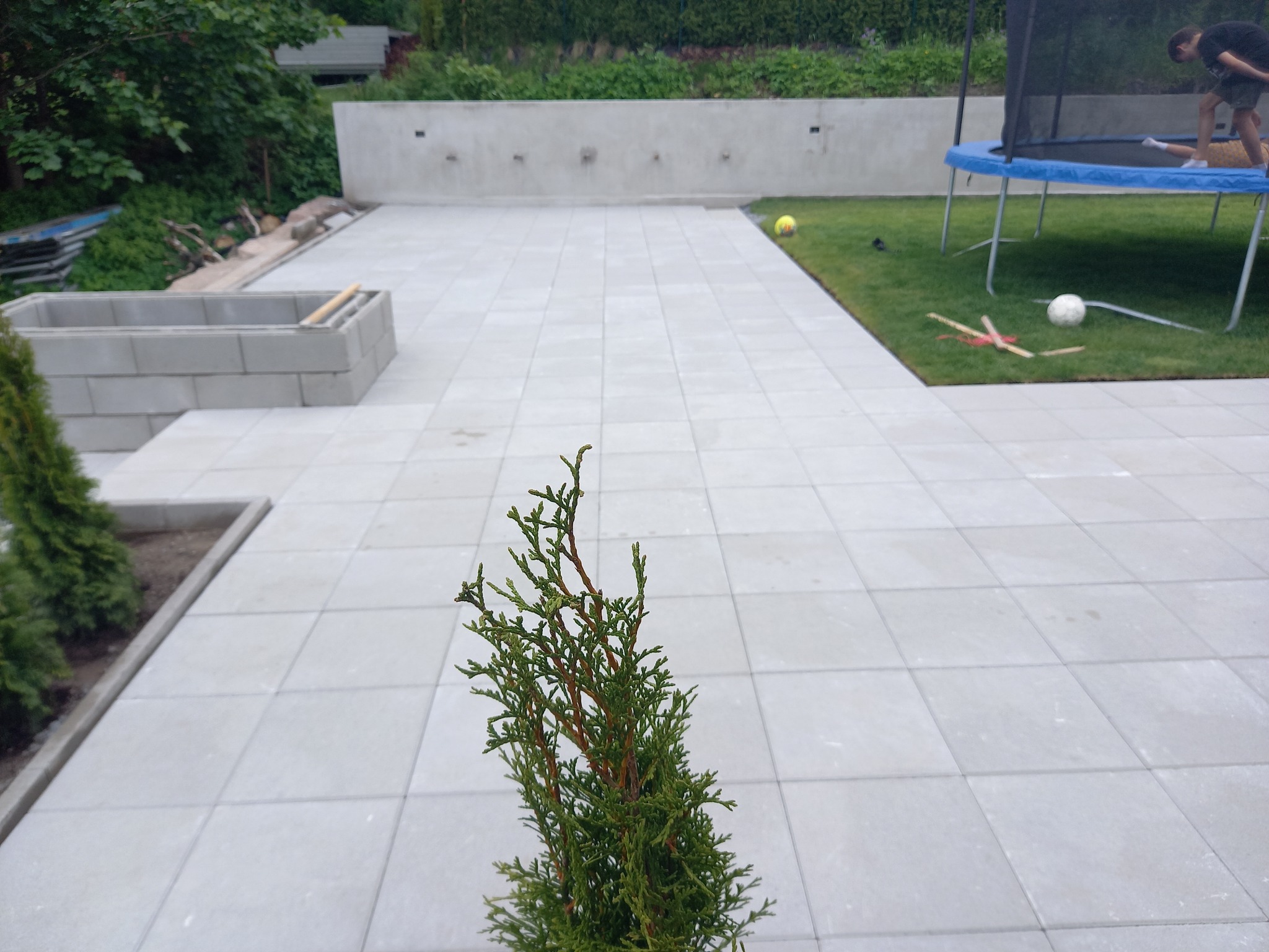 Nowoczesny taras z jasnoszarych płyt, z widoczną zielenią w tle i elementami małej architektury, takimi jak donice z bloczków betonowych i trampolina.