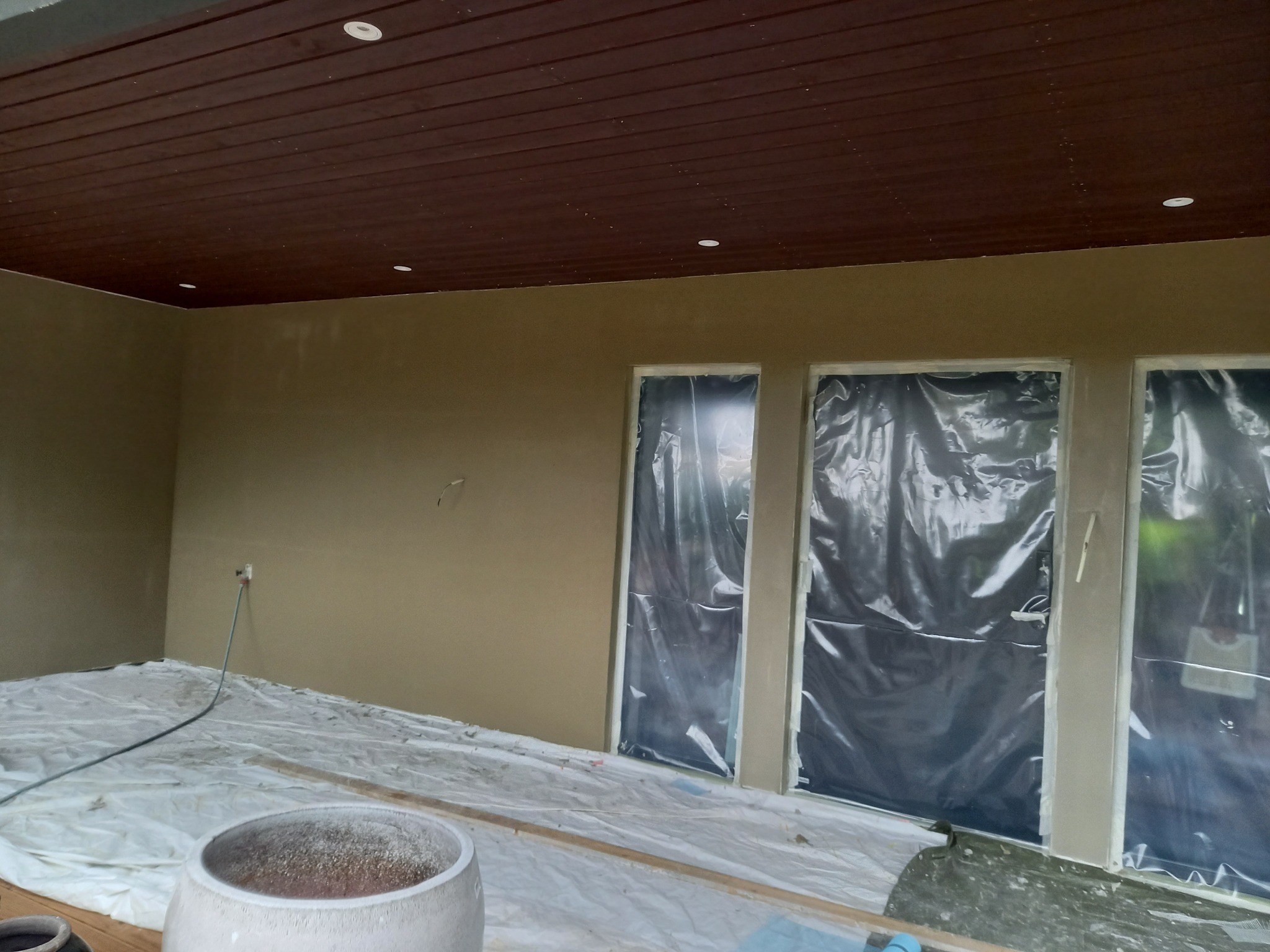 Wnętrze w trakcie prac wykończeniowych: ściany pokryte gładzią, okna zabezpieczone folią, podłoga przykryta folią ochronną. Widoczny brązowy sufit z oświetleniem.