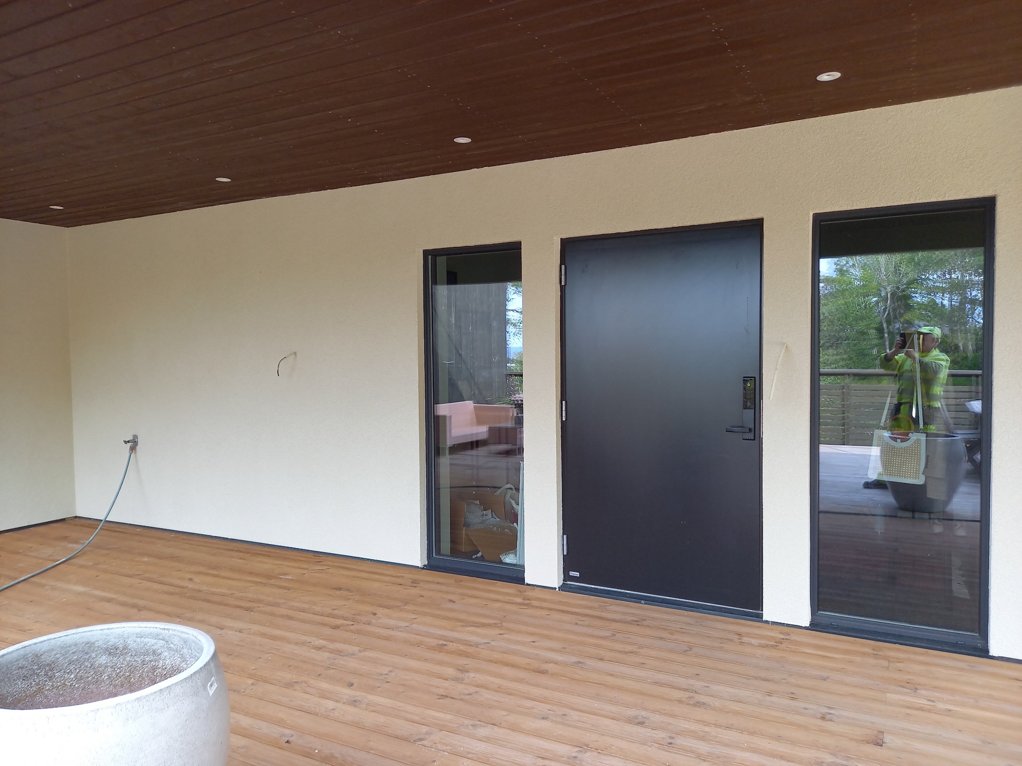 Elewacja budynku po tynkowaniu z drewnianą podłogą tarasu, czarnymi ramami okiennymi i drzwiami. Widoczny fragment wnętrza przez okno.