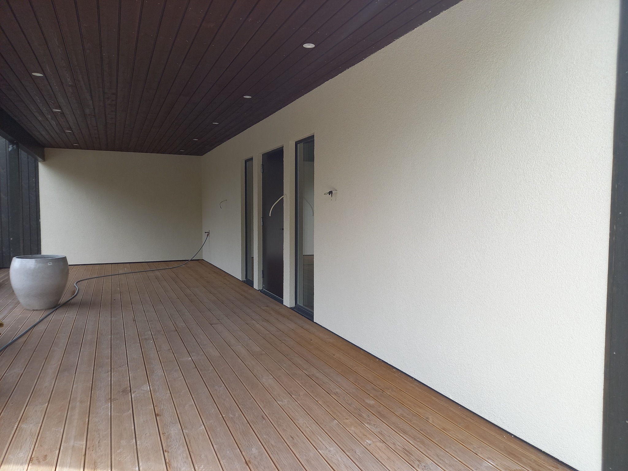 Jasna elewacja budynku z drewnianym tarasem i sufitem. Widoczne okna i donica. Teksturowana ściana, oświetlenie punktowe w suficie.