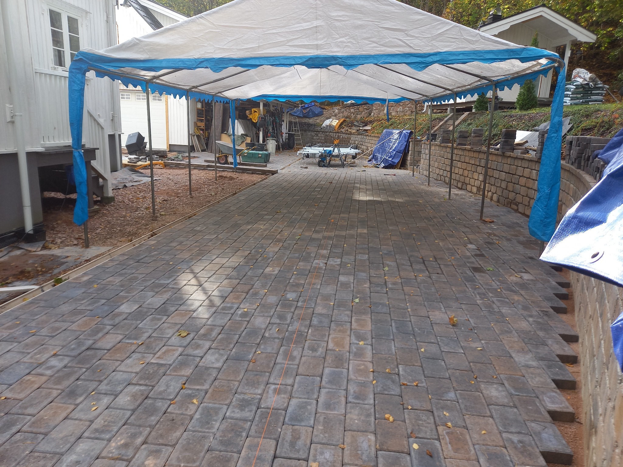 Ułożona kostka brukowa na podjeździe, widok z góry. Nad terenem rozpięty biało-niebieski namiot. W tle budynek mieszkalny i narzędzia budowlane.