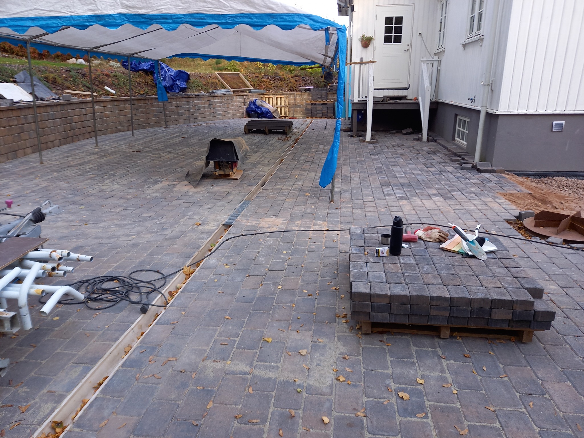 Układanie kostki brukowej na posesji domu jednorodzinnego. Widoczne narzędzia, materiały i fragmenty instalacji. Prace w toku, częściowo zadaszone.