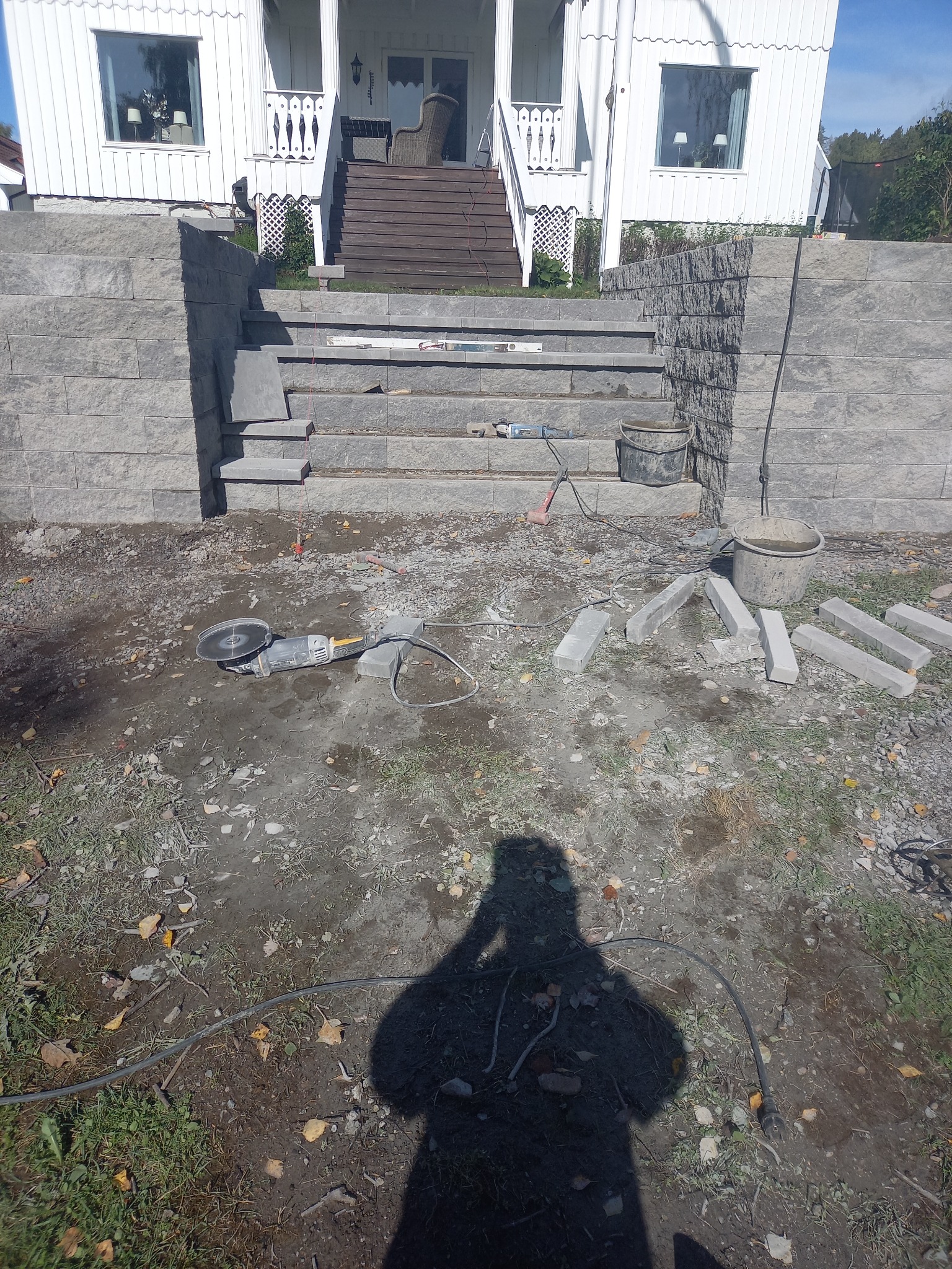 Murowane schody wejściowe do domu z szarej kostki, w trakcie budowy. Widoczne narzędzia i materiały budowlane. Cień osoby robiącej zdjęcie.