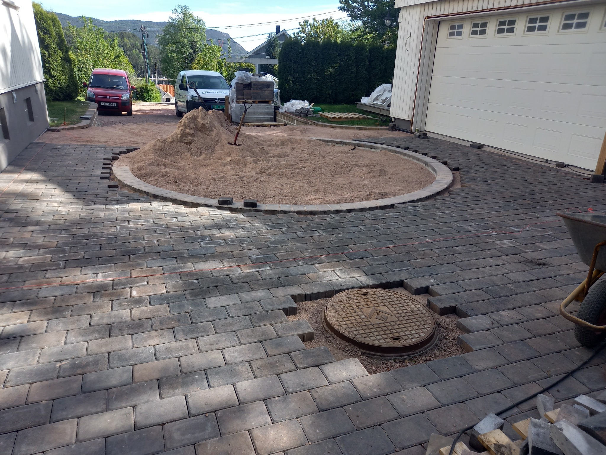 Układanie kostki brukowej na podjeździe z okrągłym klombem. Widoczny manhole, piasek, częściowo ułożona kostka i narzędzia. Prace w toku, etap przygotowania podłoża.