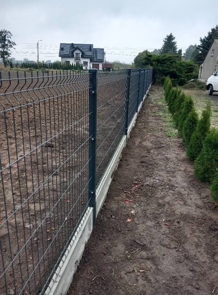 Nowoczesne ogrodzenie panelowe z podmurówką betonową, widok z boku, w tle dom jednorodzinny i rząd małych tuj. Szara, pochmurna pogoda.