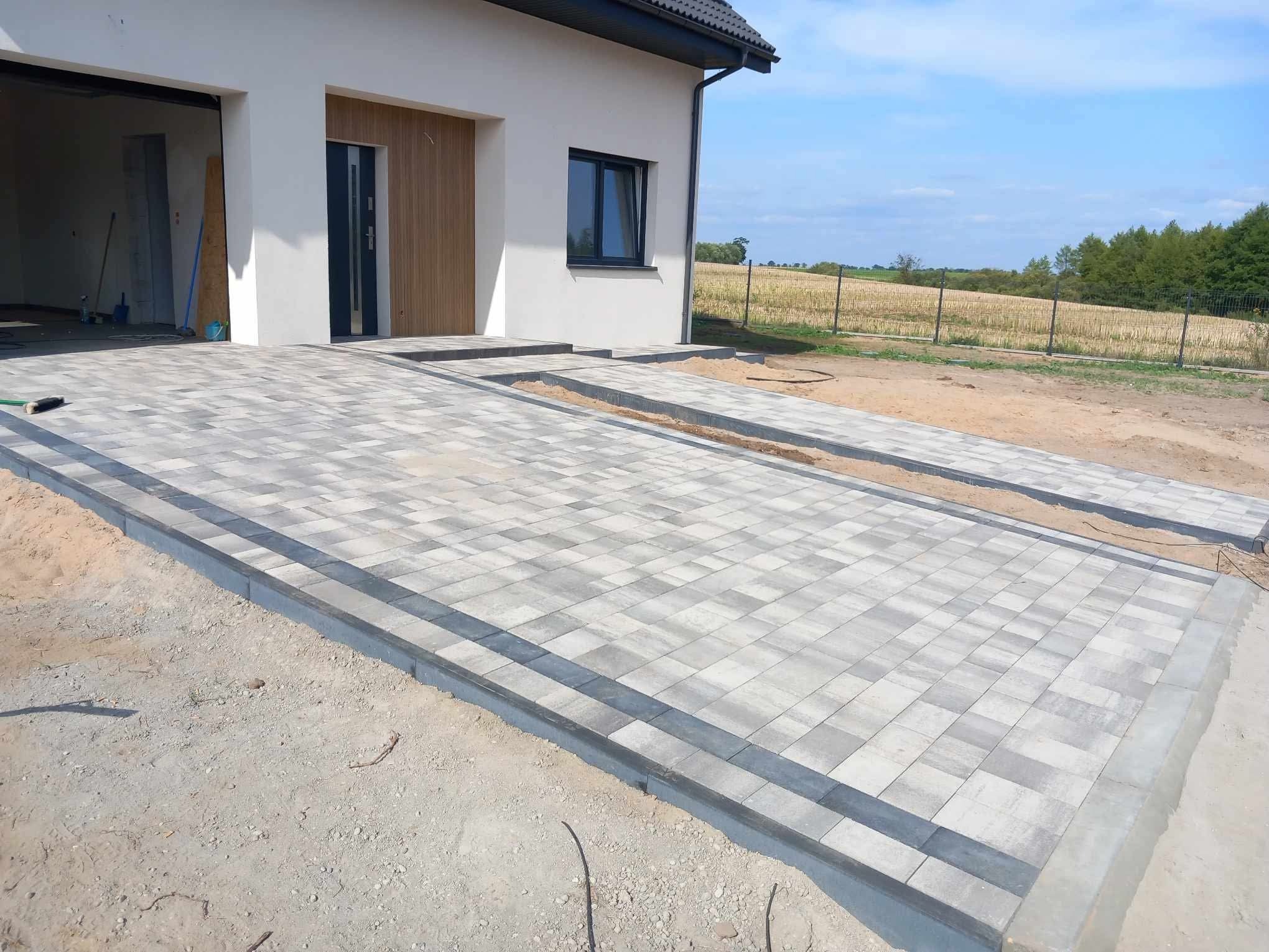 Nowoczesny podjazd z jasnoszarej kostki brukowej, obrzeża z ciemniejszej kostki. W tle dom z drewnianą elewacją i brama garażowa w stanie surowym.