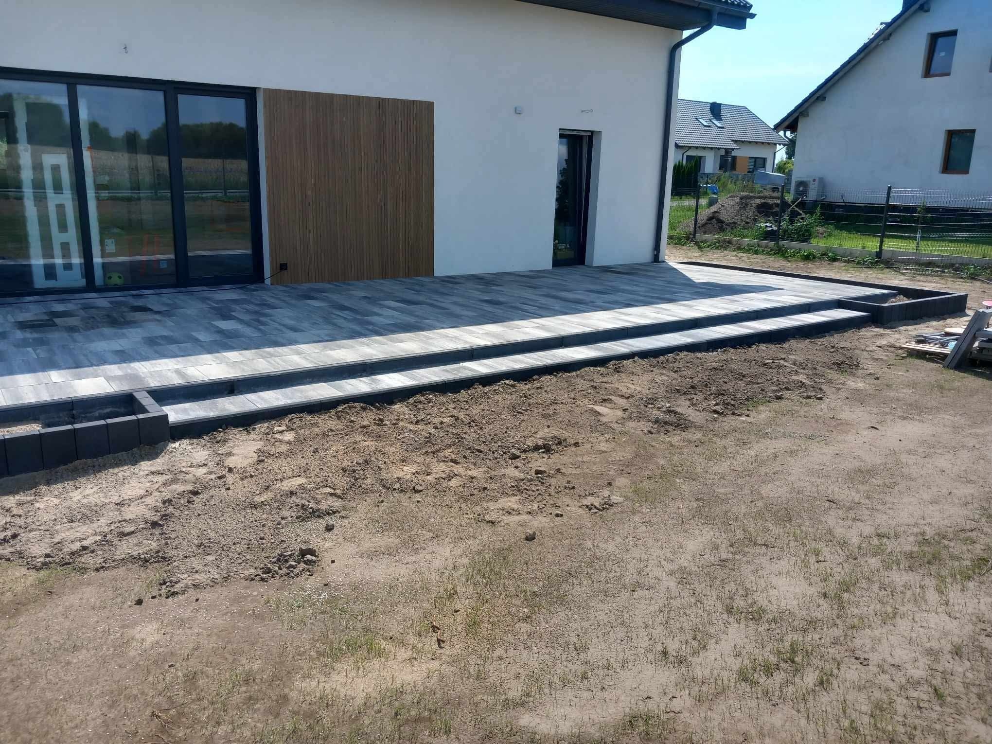 Nowoczesny taras z kostki brukowej w odcieniach szarości, z dwoma stopniami i obrzeżami, przylegający do białego budynku z drewnianą elewacją. Widok na ogród i sąsiednie domy.