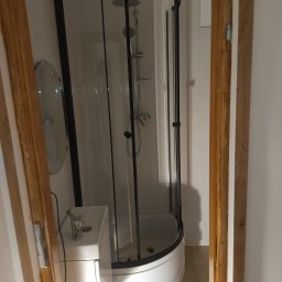 Nowoczesna kabina prysznicowa z czarnymi profilami, umywalka z szafką i okrągłe lustro w wykończonej łazience, widok przez drewnianą futrynę drzwi.