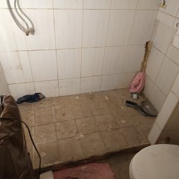 Łazienka w trakcie demontażu płytek, widoczny fragment toalety, ślady po kleju, narzędzia i śmieci. Przygotowanie do remontu.