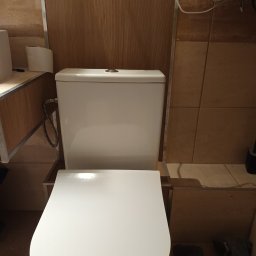 Biała toaleta z deską w brązowej łazience z beżowymi płytkami. Widoczne elementy instalacji i zabudowy. Ujęcie z góry, bliskie.