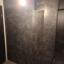 Arian Wolwiak AS-Budowa - Nowoczesna zabudowa ściany z szarych paneli z efektem betonu, tworząca szafę wnękową z półkami. Widoczne oświetlenie sufitowe i fragment podłogi z płytek.