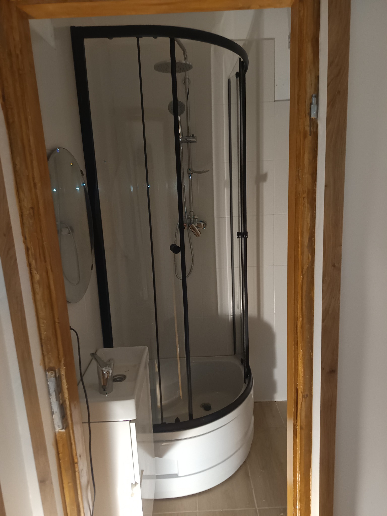 Nowoczesna kabina prysznicowa z czarnymi profilami, umywalka z szafką i okrągłe lustro w wykończonej łazience, widok przez drewnianą futrynę drzwi.