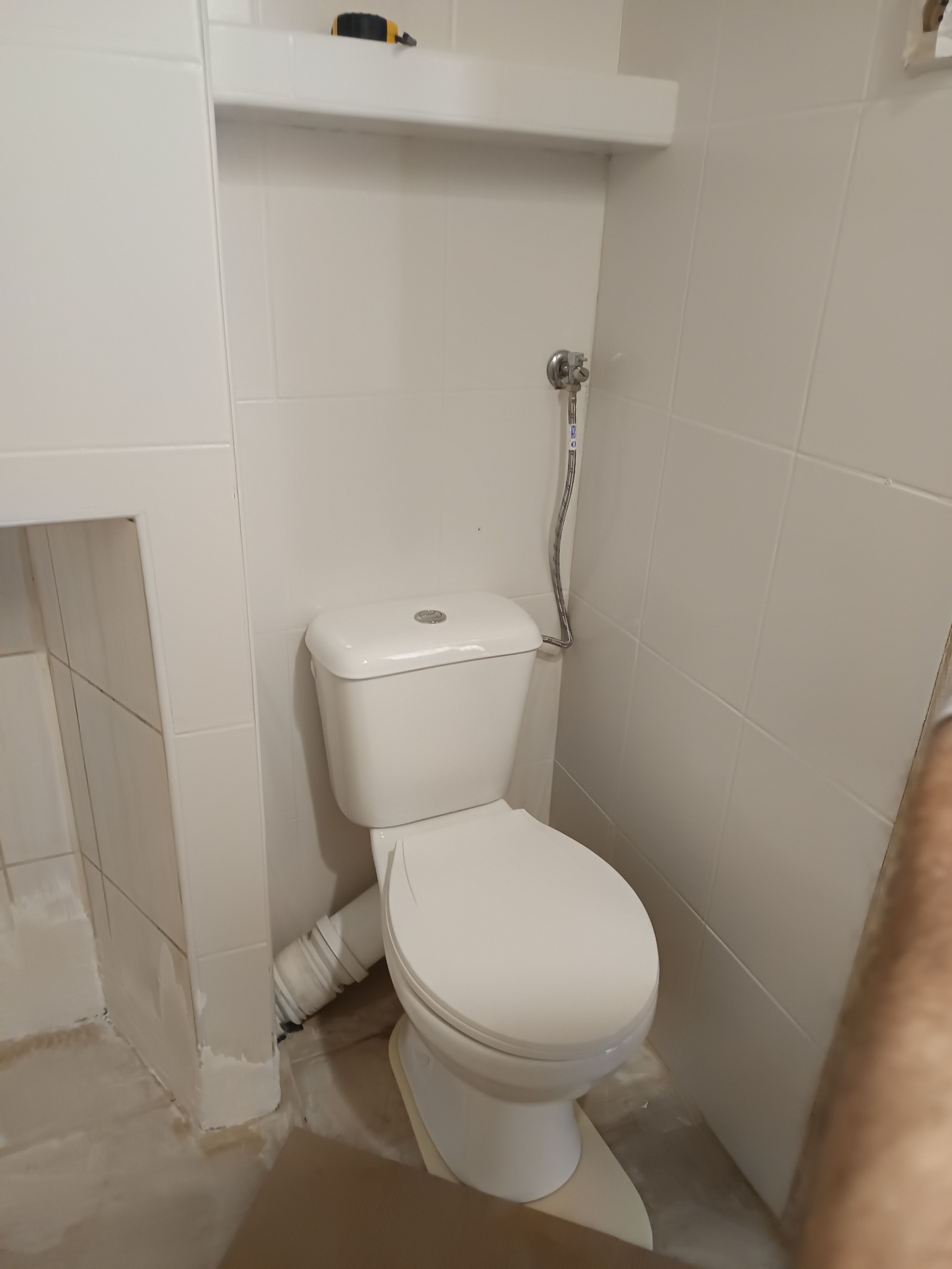 Biała toaleta kompaktowa w wykończonej, jasnej łazience z płytkami. Widoczna instalacja wodna i półka nad toaletą. Ujęcie z bliska, z boku.