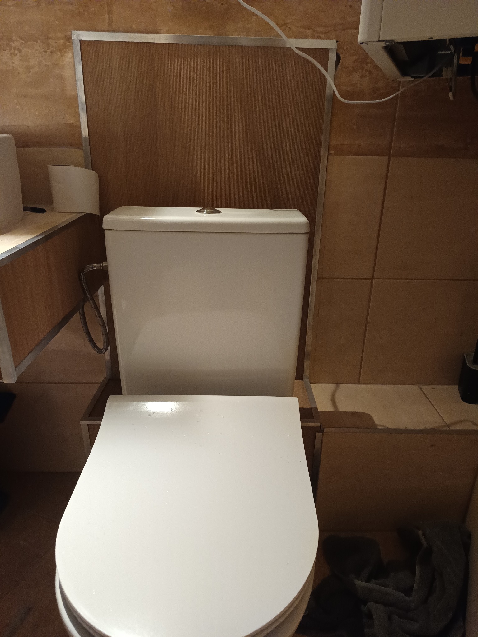 Biała toaleta z deską w brązowej łazience z beżowymi płytkami. Widoczne elementy instalacji i zabudowy. Ujęcie z góry, bliskie.