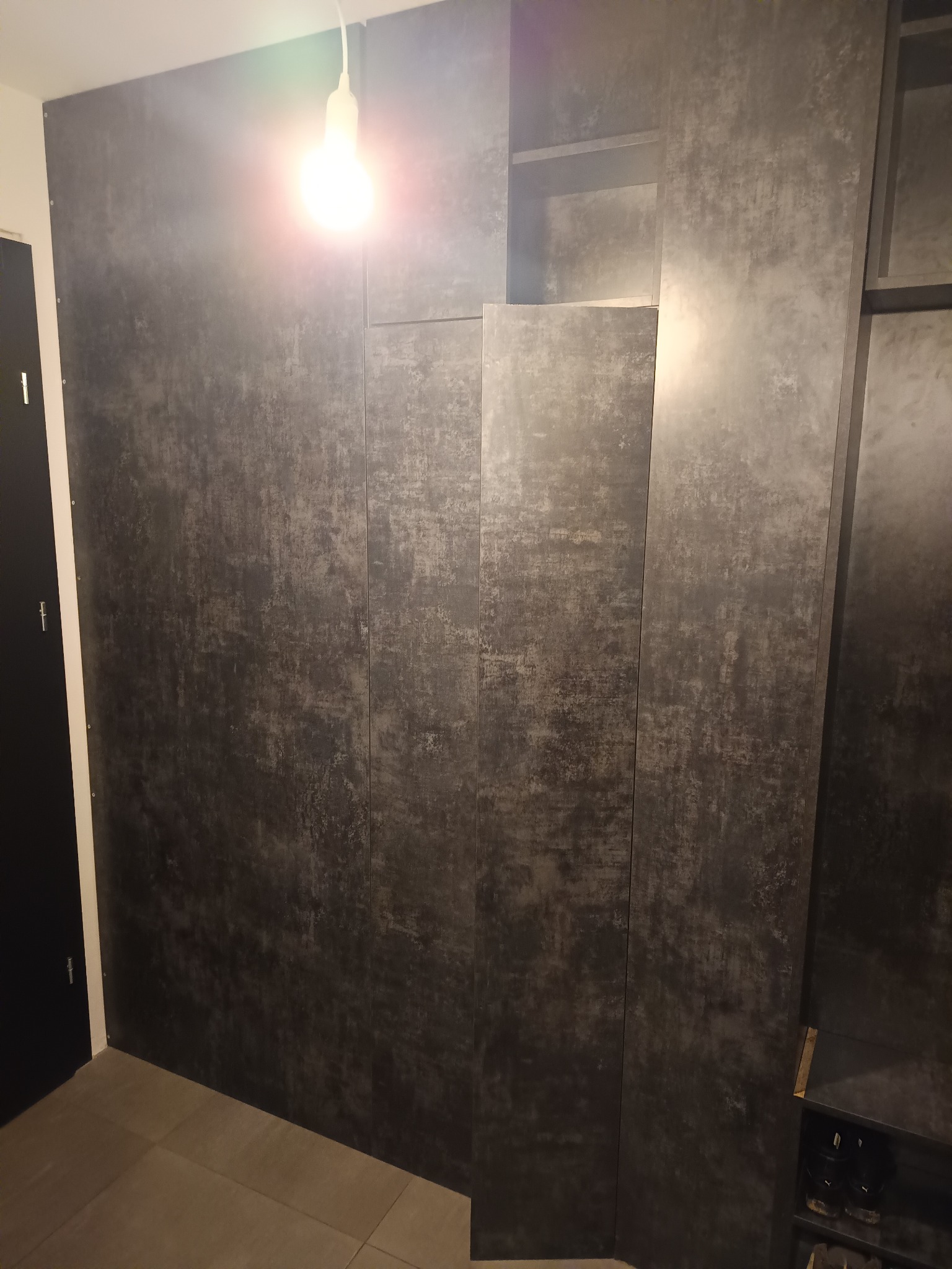 Nowoczesna zabudowa ściany z szarych paneli z efektem betonu, tworząca szafę wnękową z półkami. Widoczne oświetlenie sufitowe i fragment podłogi z płytek.