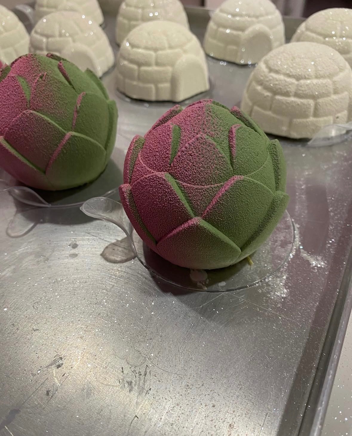 Artystyczne desery: dwa musowe kwiaty w odcieniach zieleni i różu na srebrnej tacy, w tle igloo z białej czekolady. Unikalna kompozycja i tekstura.