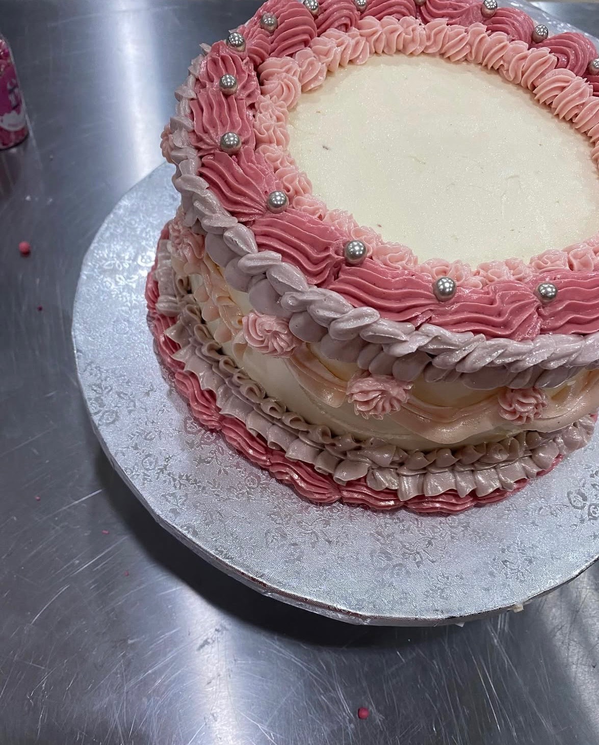 Dekorowany tort z kremem w odcieniach różu i srebrnymi kuleczkami, umieszczony na srebrnej paterze. Widok z góry, ukazujący detale zdobień.