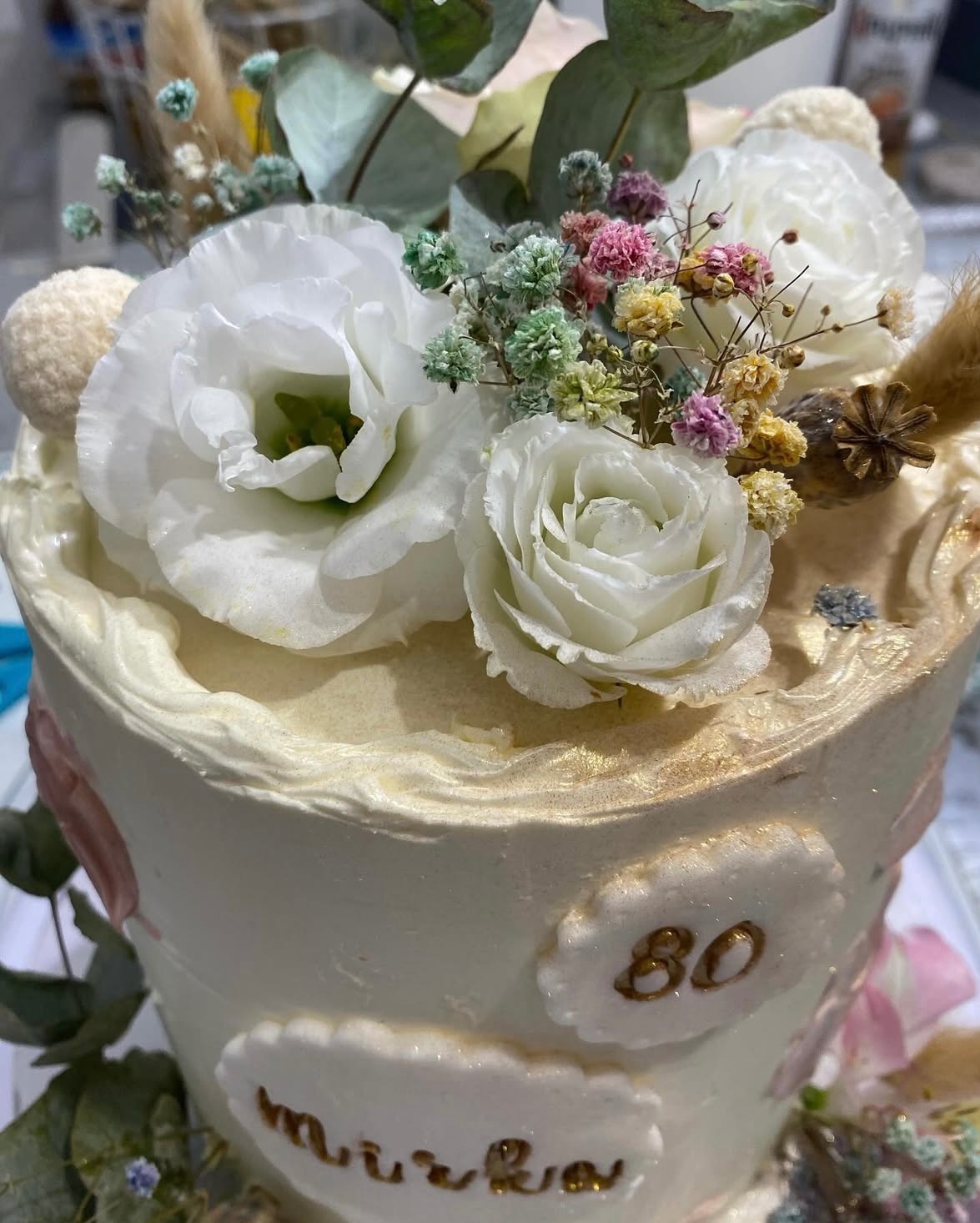 Tort urodzinowy z białym kremem, kwiatami i napisem 'Mirka' oraz cyfrą '80' w złotym kolorze. Dekoracja z suszonych roślin i kulek.