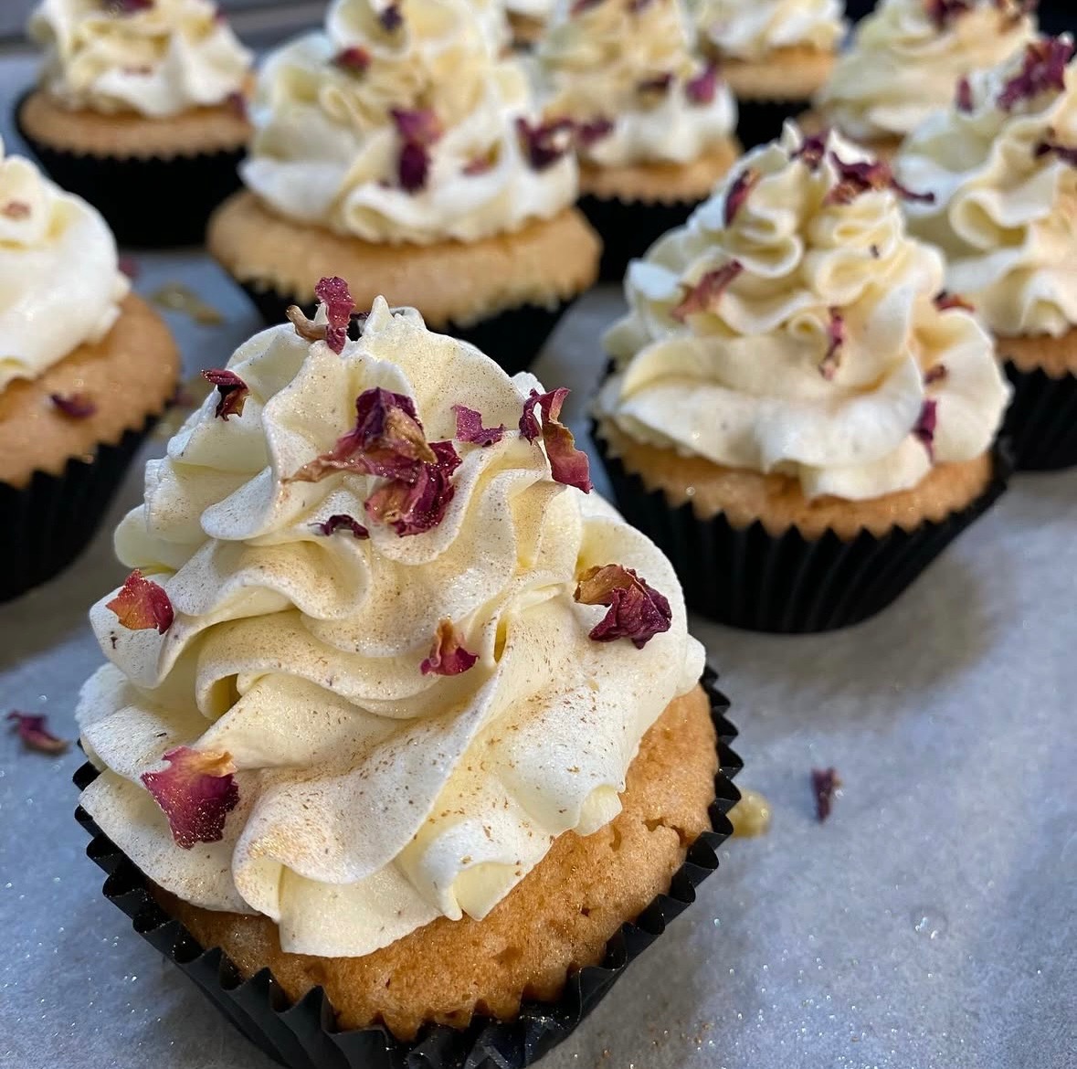 Delikatne babeczki z kremem i płatkami róż w czarnych papilotkach, ustawione w rzędzie. Krem ma subtelny, waniliowy odcień, a płatki dodają elegancji.