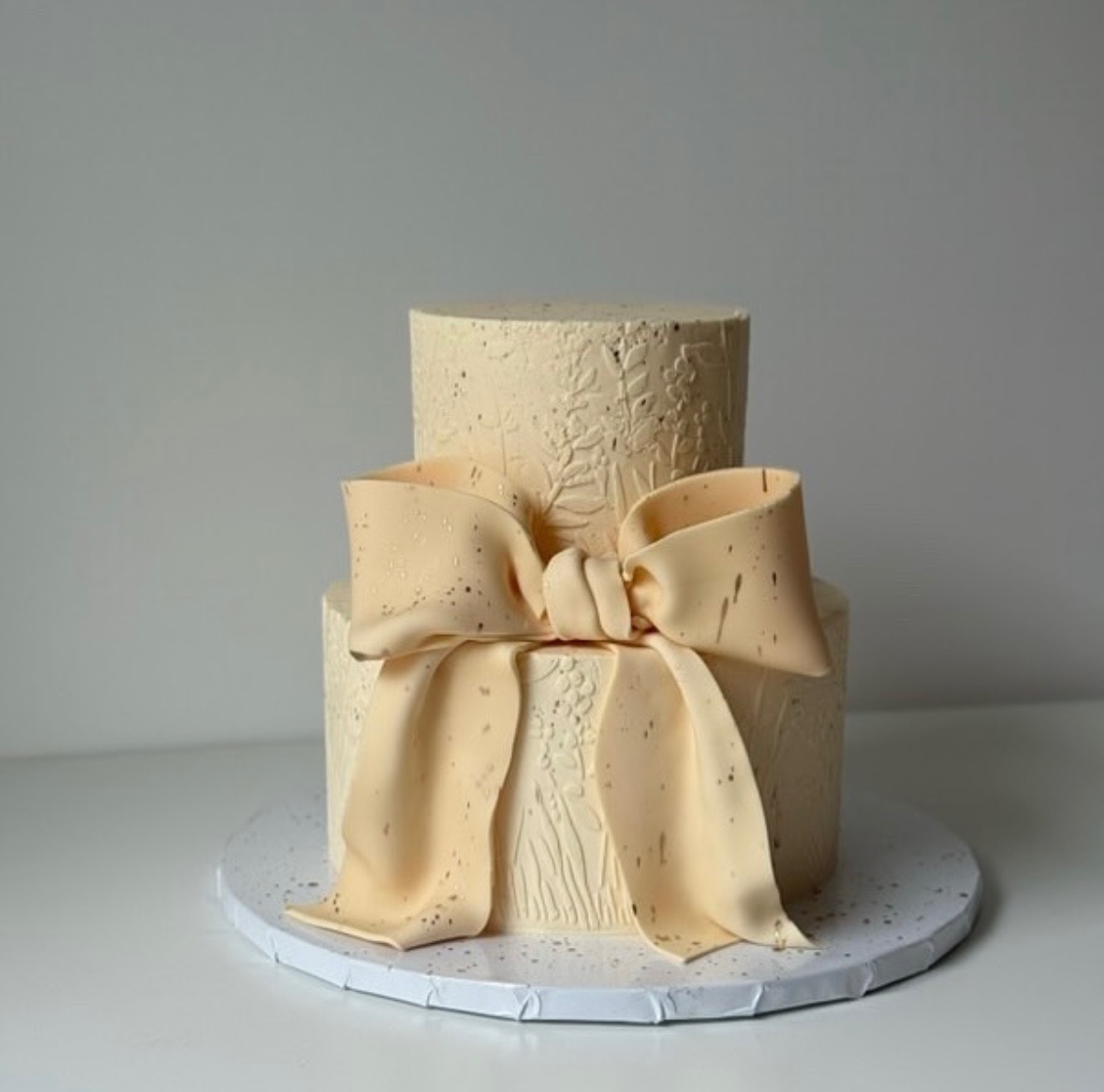 Dwupoziomowy tort w kremowym odcieniu, zdobiony wytłaczanym wzorem roślinnym i dużym, satynowym kokardą z delikatnymi, złotymi akcentami.