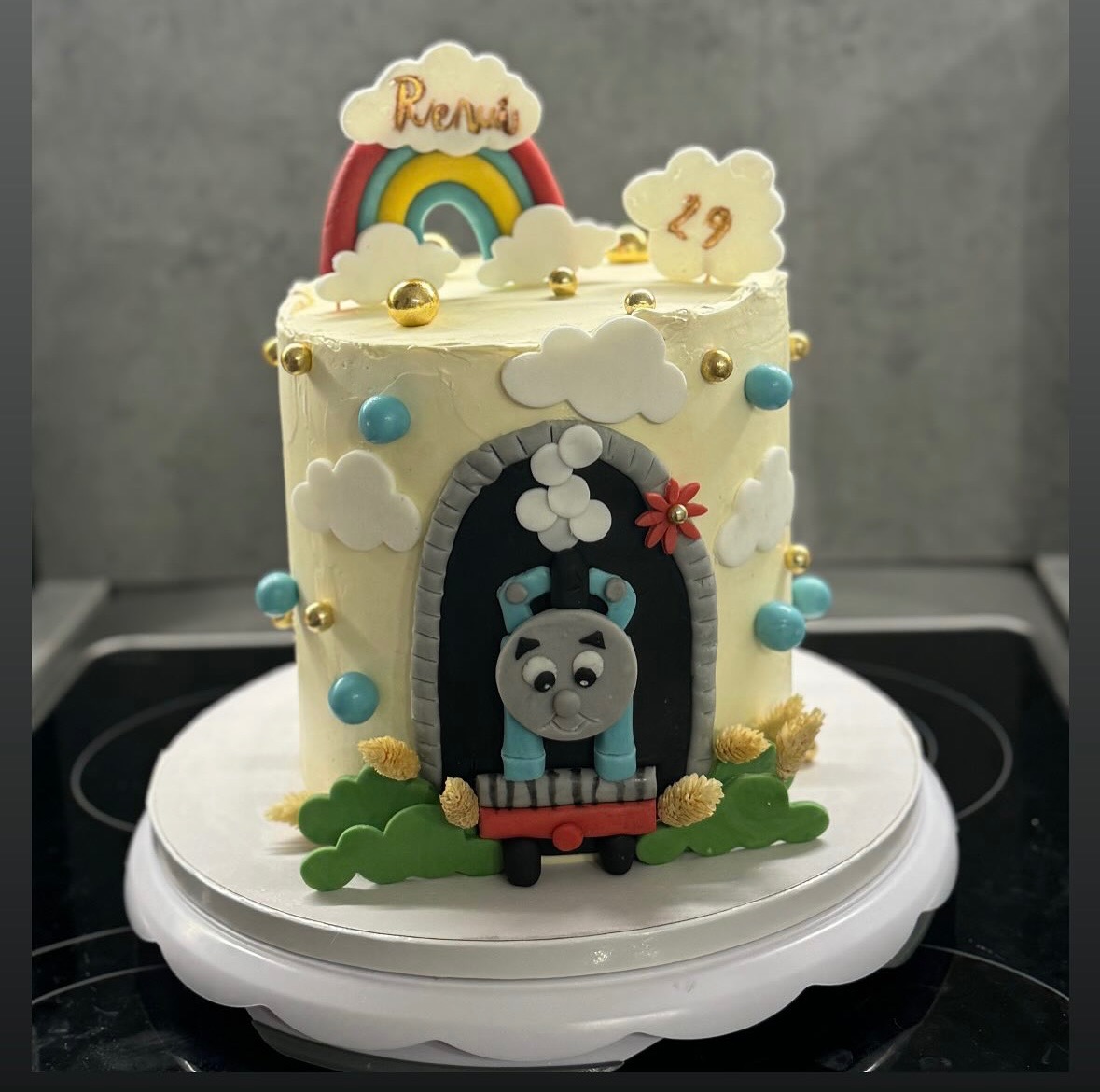 Tort urodzinowy z motywem Tomka i Przyjaciół, dekorowany tęczą, chmurkami i napisem 'Rena', stojący na białym talerzu obrotowym.