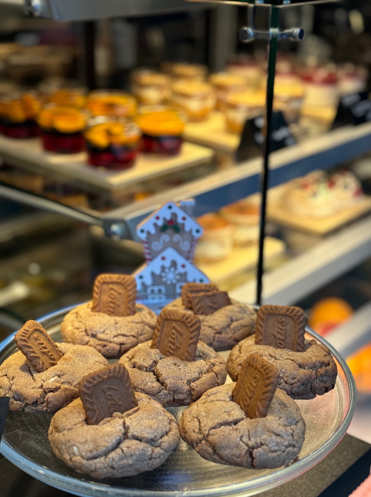 Ciasteczka z ciasteczkiem Lotus na srebrnej tacy, w tle inne desery w witrynie cukierni. Dekoracja świąteczna w postaci piernikowego domku.