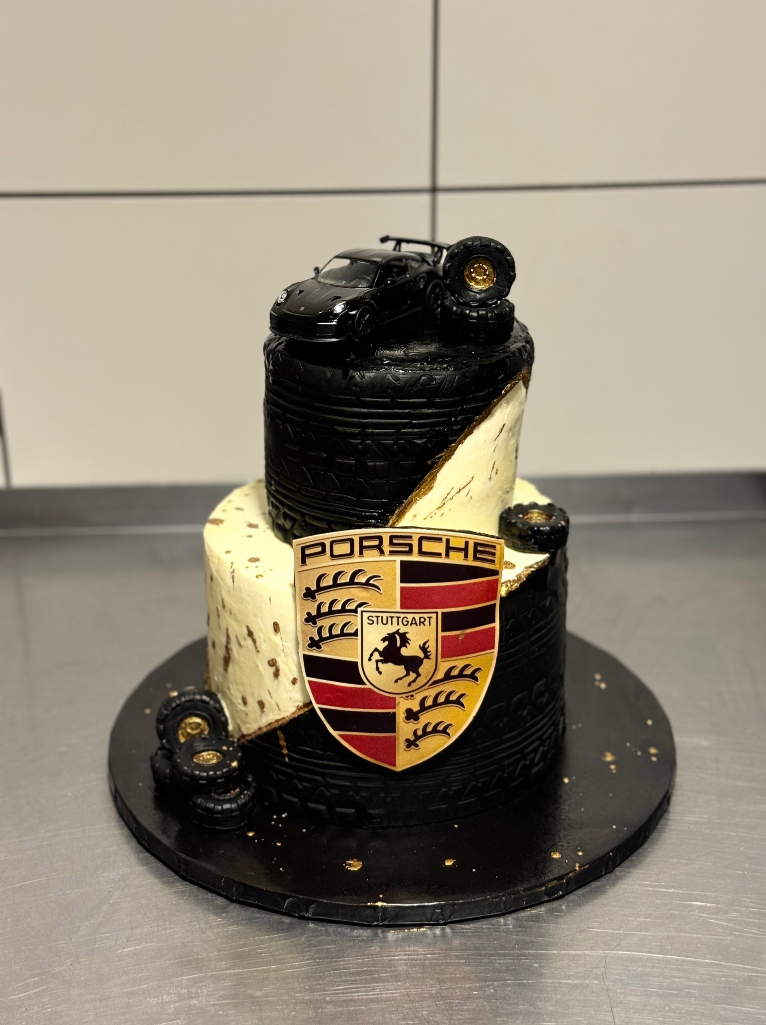 Dwupoziomowy tort z motywem Porsche: czarne opony, złote akcenty, model auta na szczycie i herb Stuttgartu. Idealny dla fana motoryzacji.